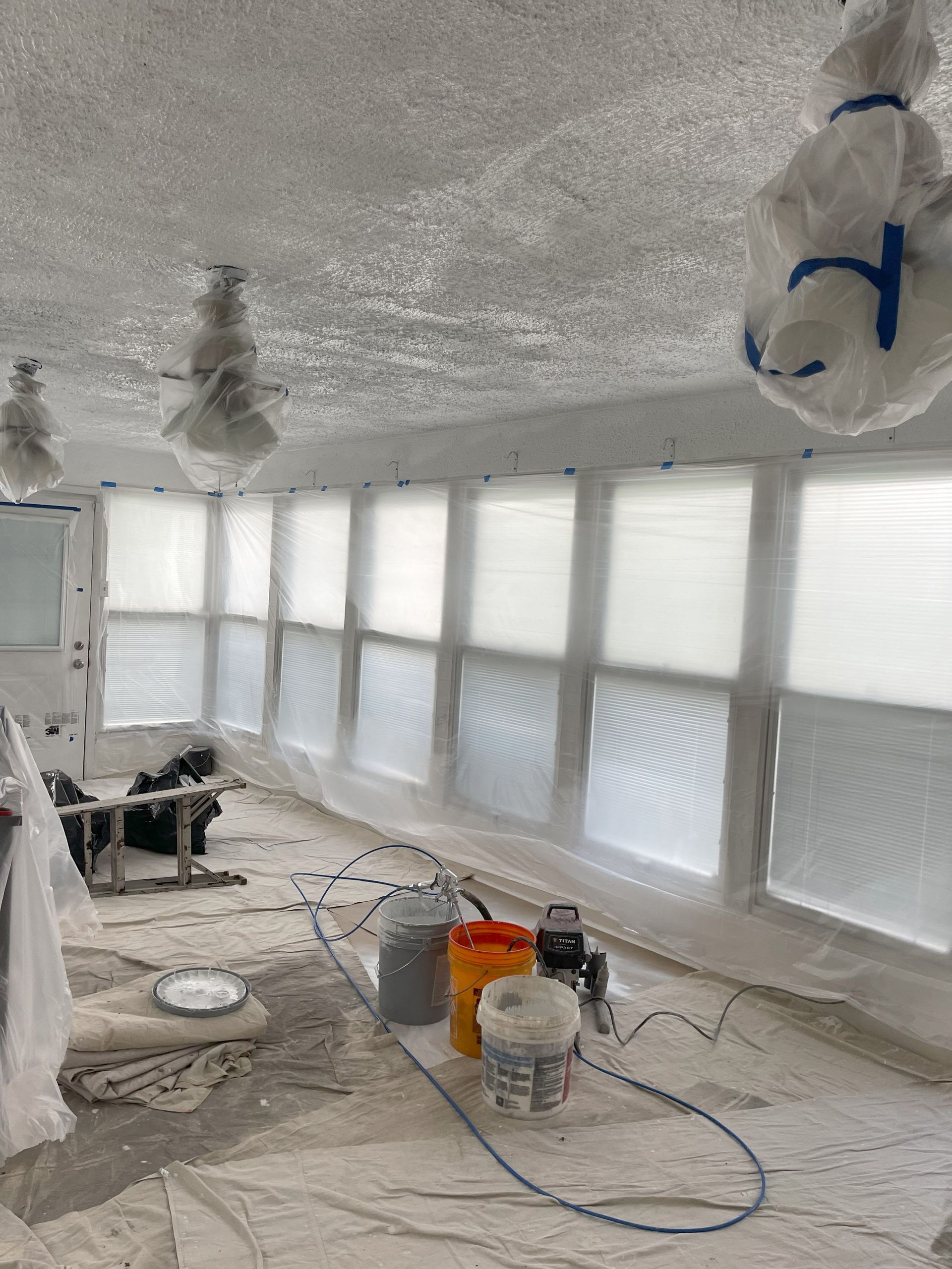 Room being painted, windows covered, ceiling and floor masked. Paint buckets and spray equipment visible.
