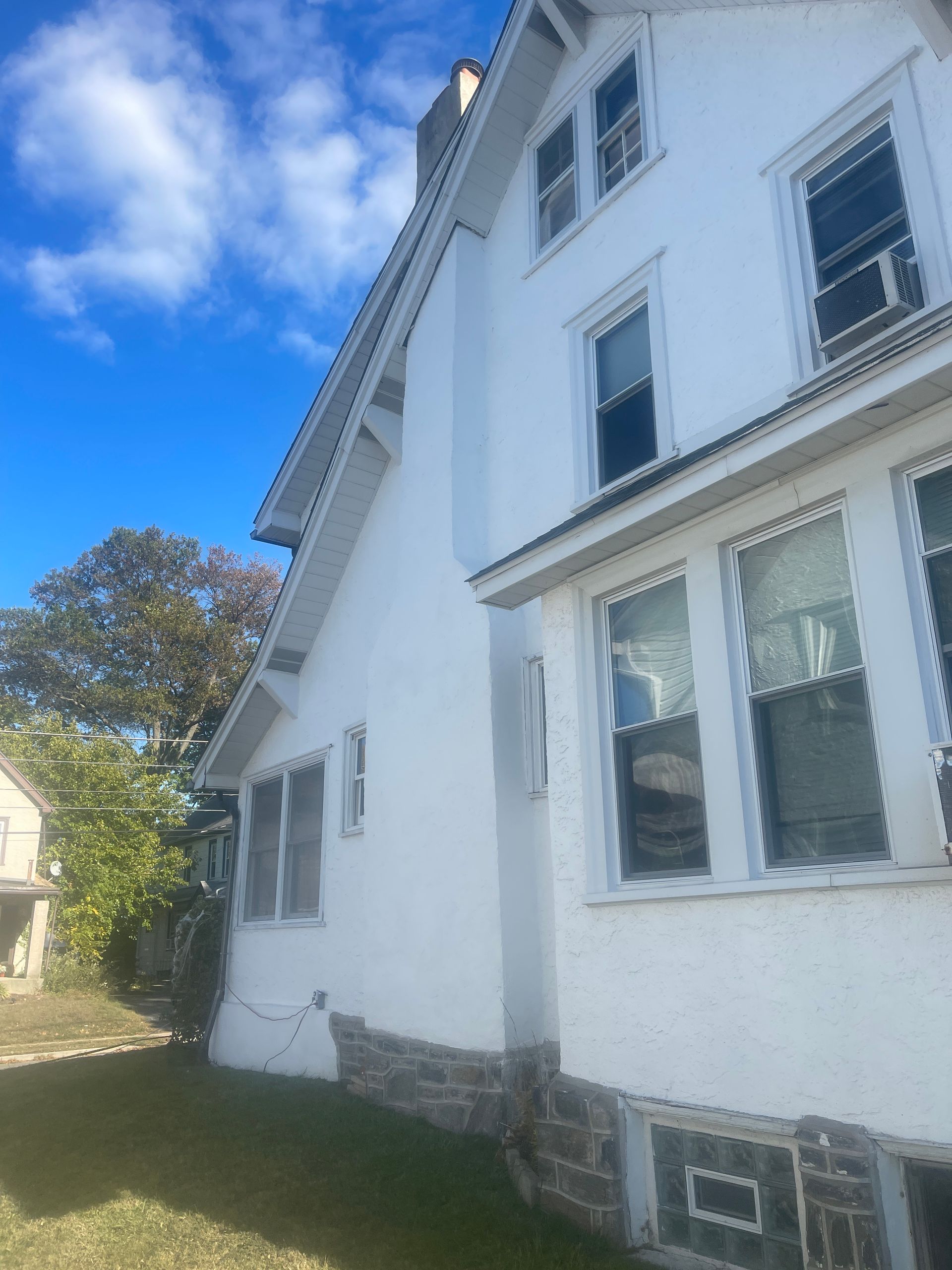 White two-story house with stone foundation. Blue sky with white clouds. Green grass and a tree are visible.