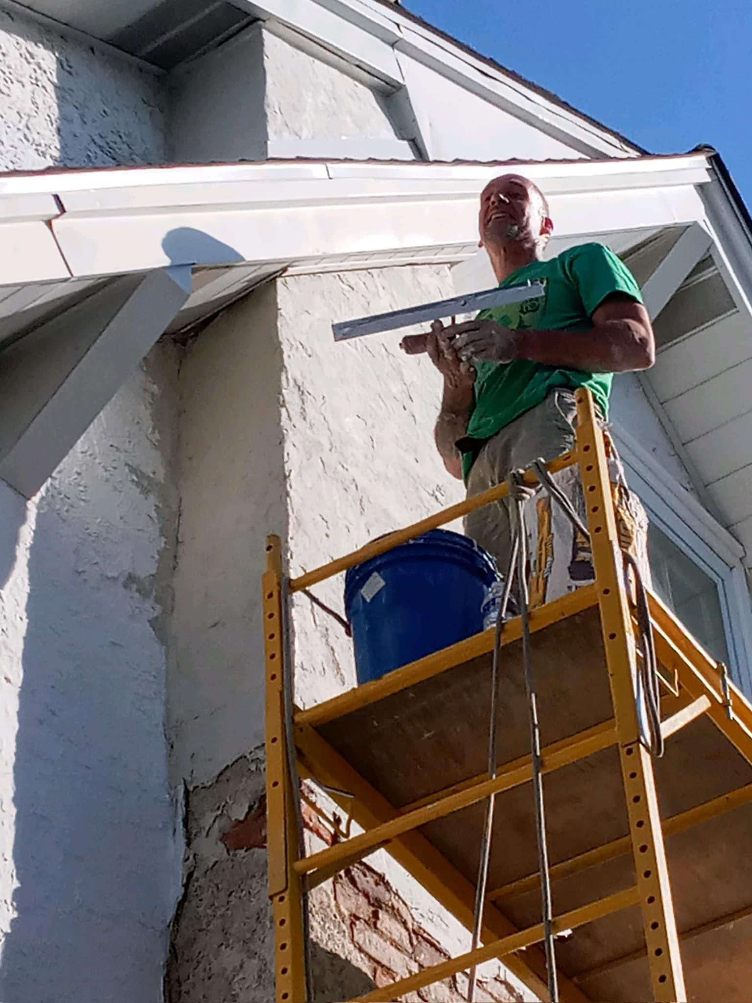 Man on scaffolding applies stucco to exterior wall. Blue bucket on scaffolding.