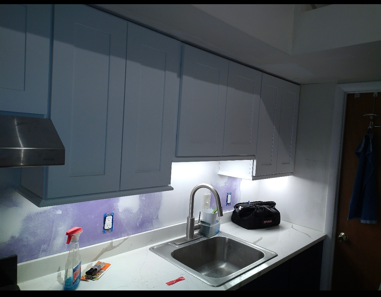 Kitchen with white cabinets, stainless steel sink, and under-cabinet lighting. Purple backsplash visible.