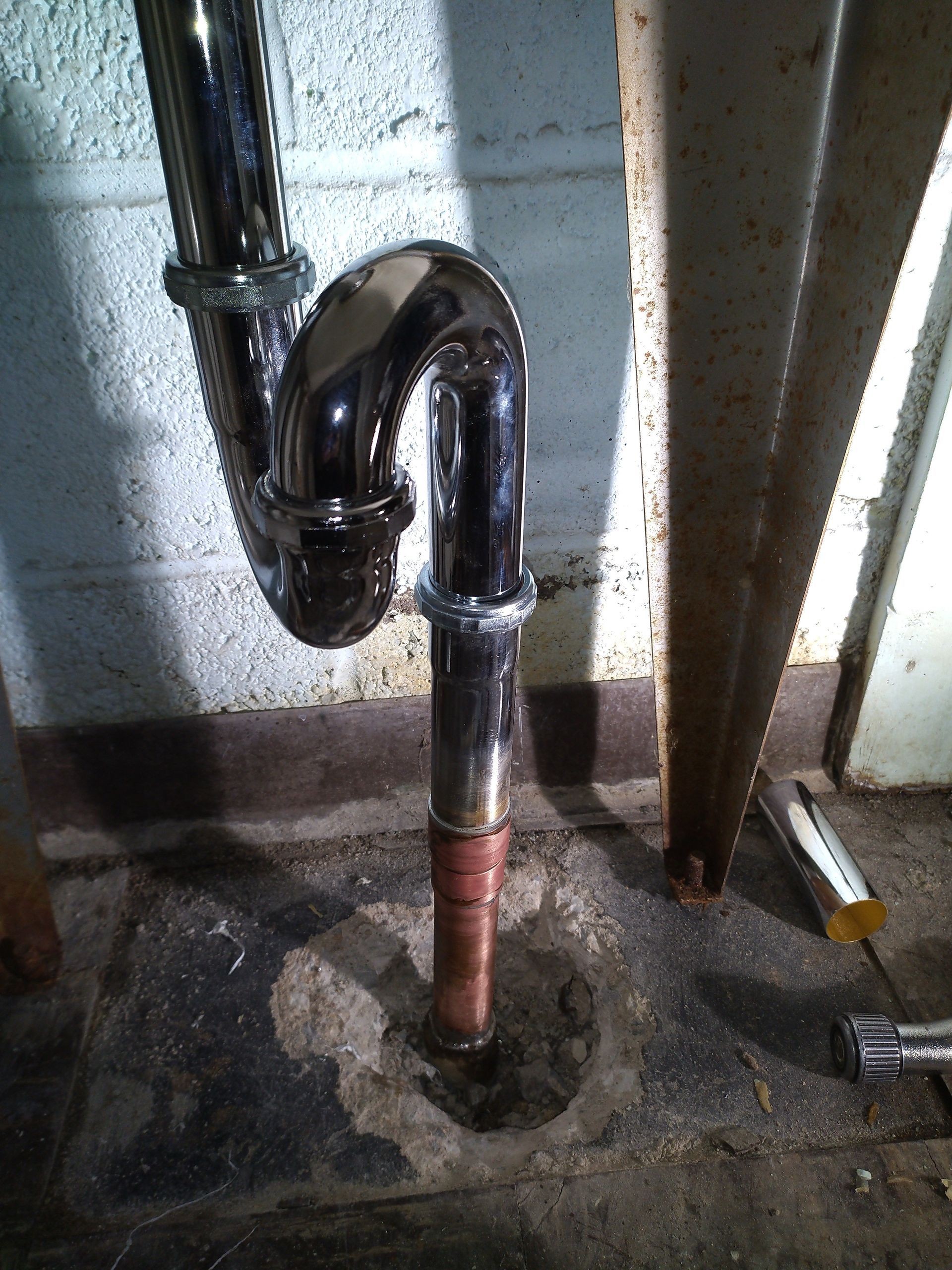 Chrome plumbing trap beneath a sink, set against a concrete wall and floor.