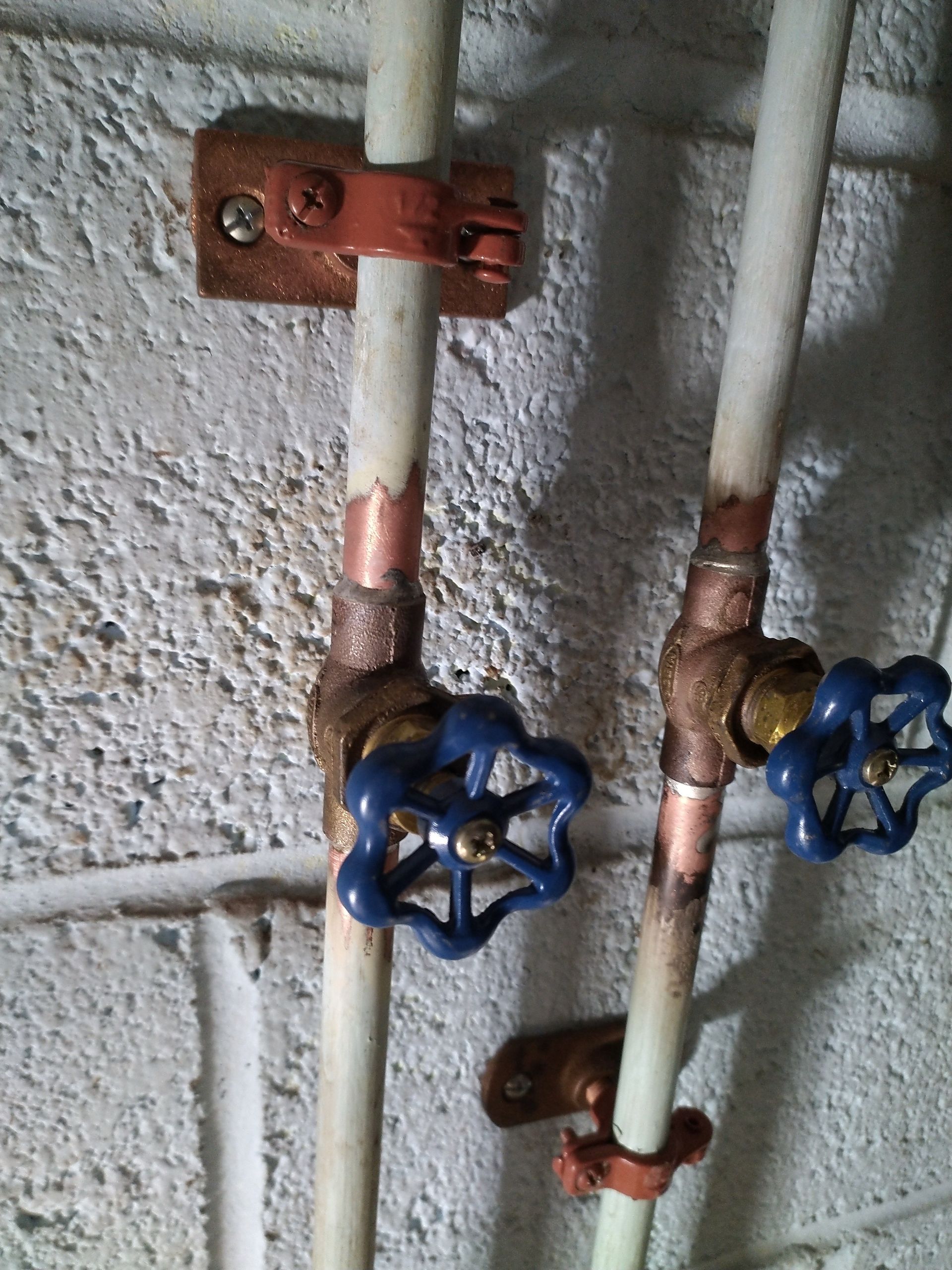 Two white water pipes with blue-handled valves, copper fittings, mounted on a brick wall.