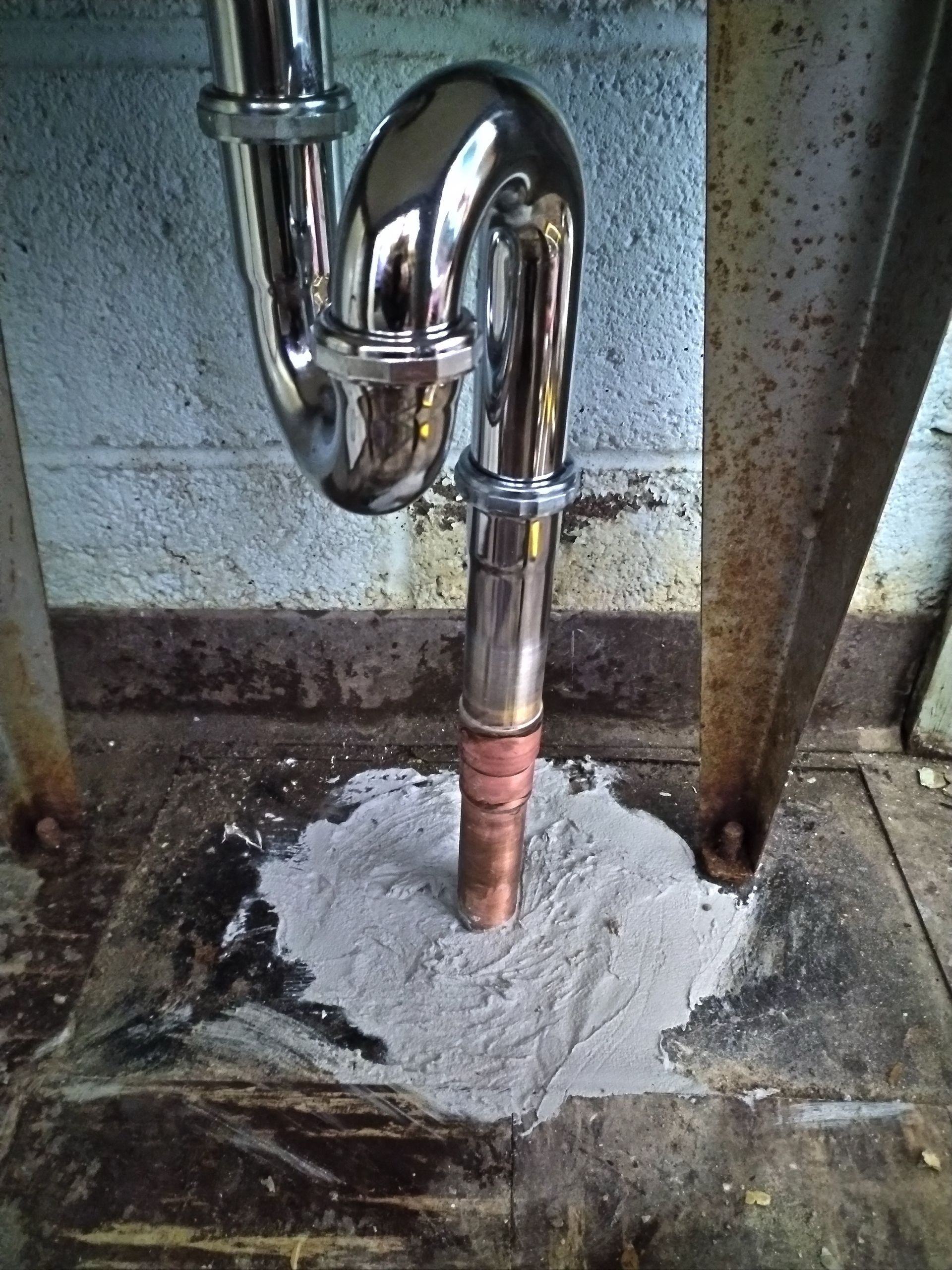 A chrome sink drain pipe is connected to a copper pipe emerging from a concrete floor.