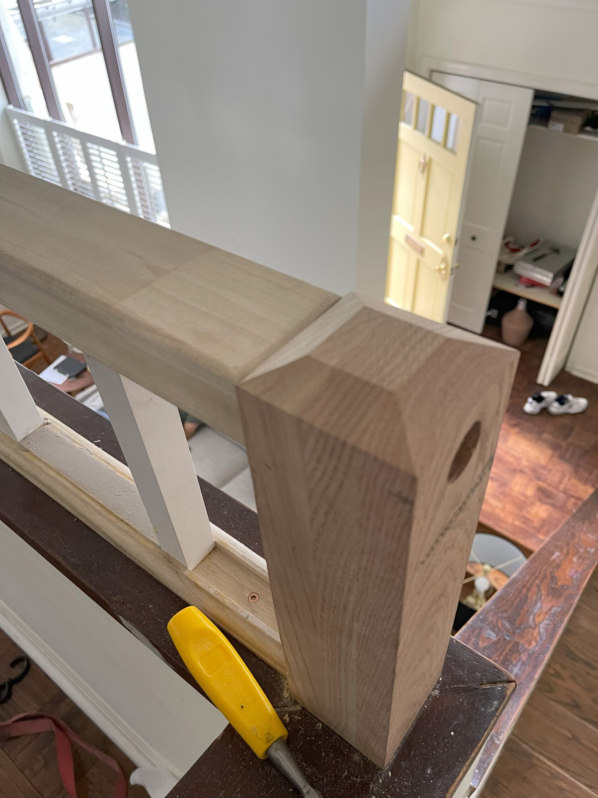 Wooden stair railing post with a hole, being worked on with a yellow tool. Interior shot.