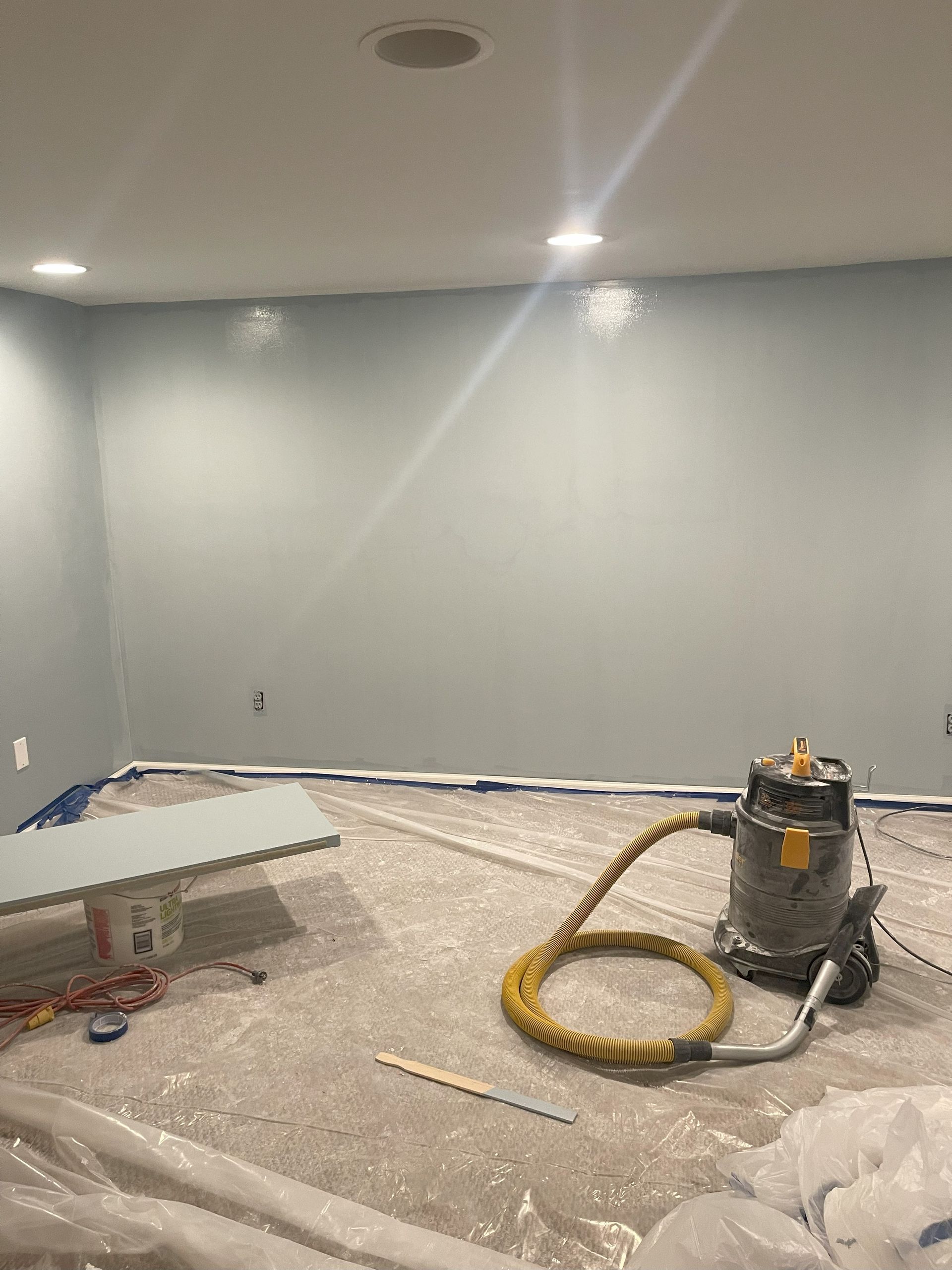 Room with light blue walls, paint sprayer, and floor covered in plastic during painting.