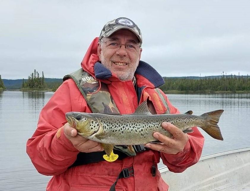 Un homme en veste rouge tient une truite brune dans ses mains.