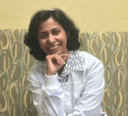 Dr. Padma Jonnavithula, BDS, DMD woman in a white shirt is sitting on a couch with her hand on her chin .