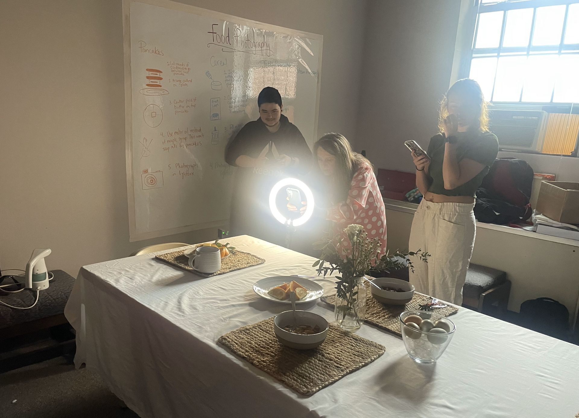 Students setting up the lighting for their food styling photos. The table is lined out with cereal, oranges, and coffee.
