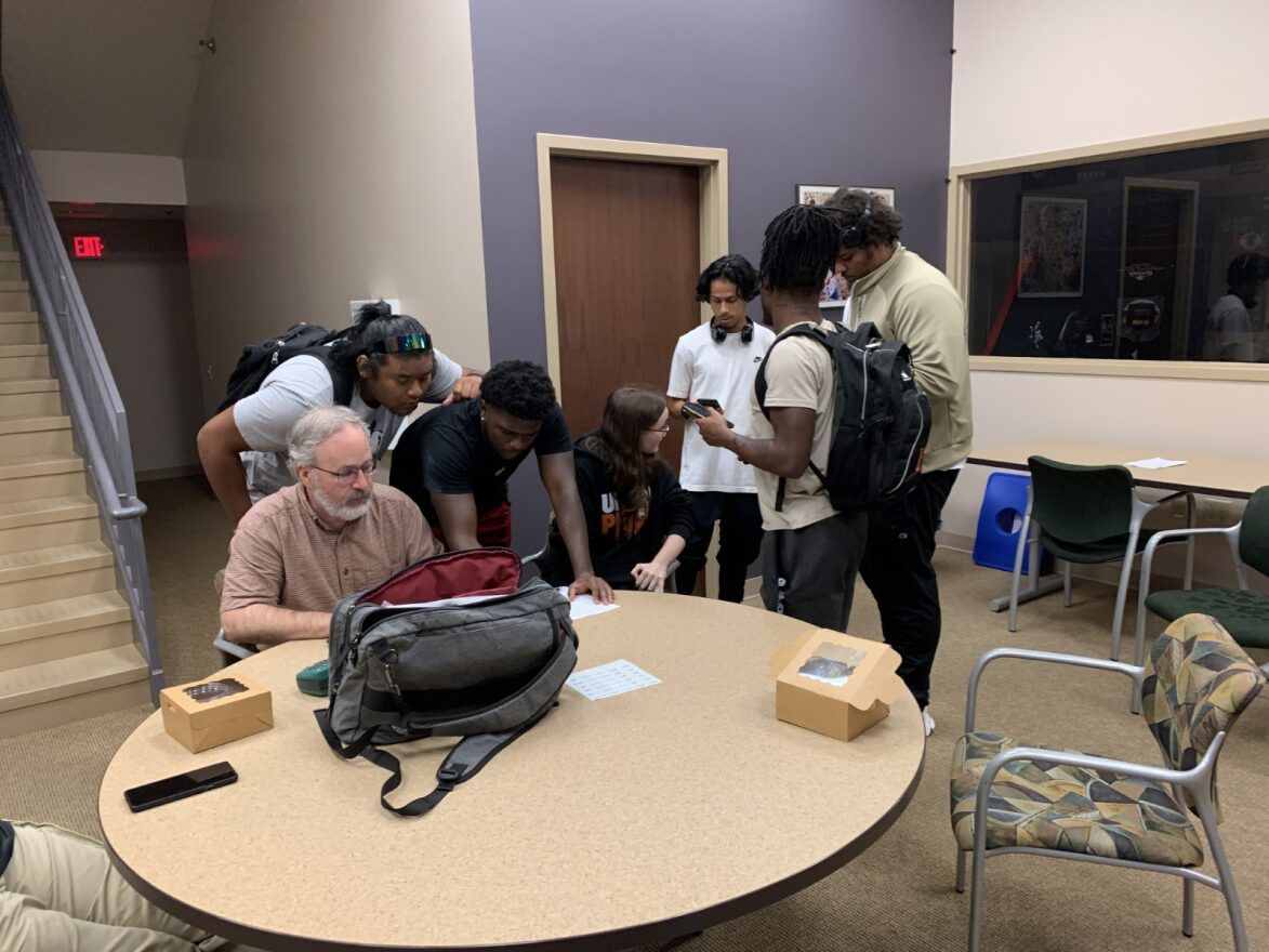 Bruce Cory sits at a table in the Black Tech lobby with members of the Esports Club surrounding him. Two students are leaning in close to Cory to read or sign a paper, while others are talking with each other close by. A gray laptop bag, phone, and food boxes are on the table.