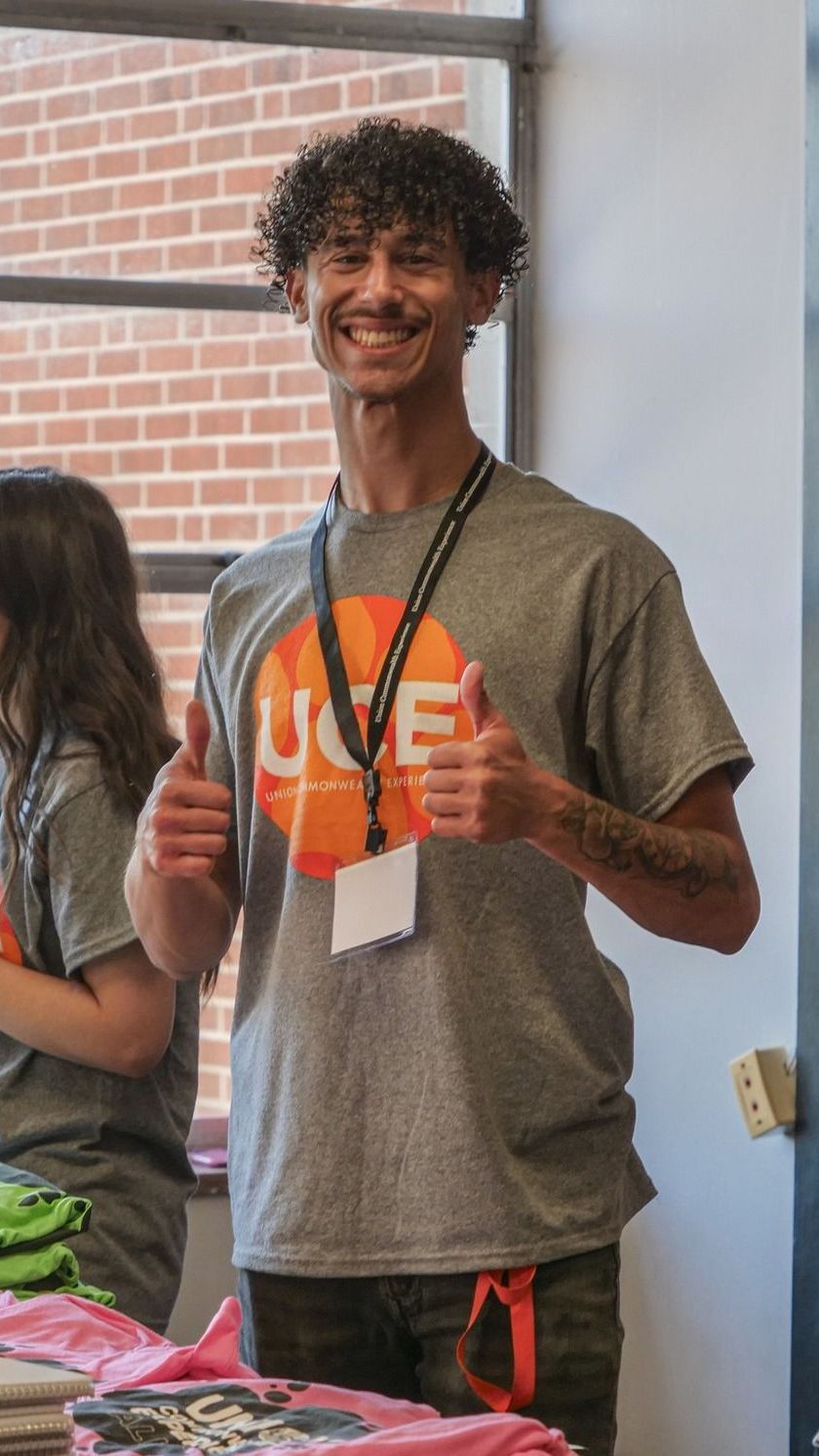 Tyce is standing behind a table with piles of Union merchandise on it. He is smiling at the camera, givign the thumbs-up sign with both hands, and wearing a lanyard and a gray shirt with an orange UCE logo on it.
