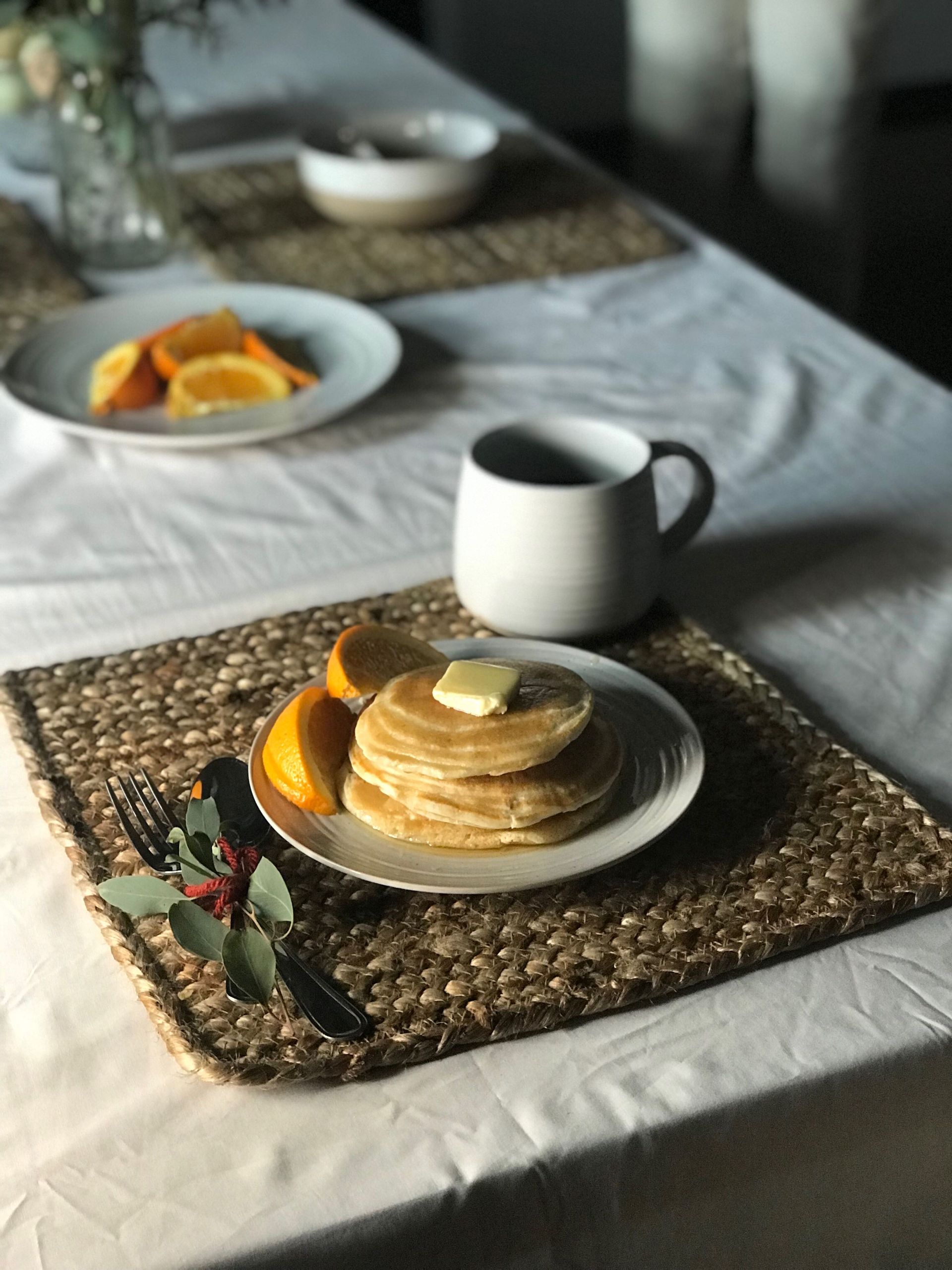 A plate of pancakes and oranges sits on a woven tray with a fork and a coffee cup.