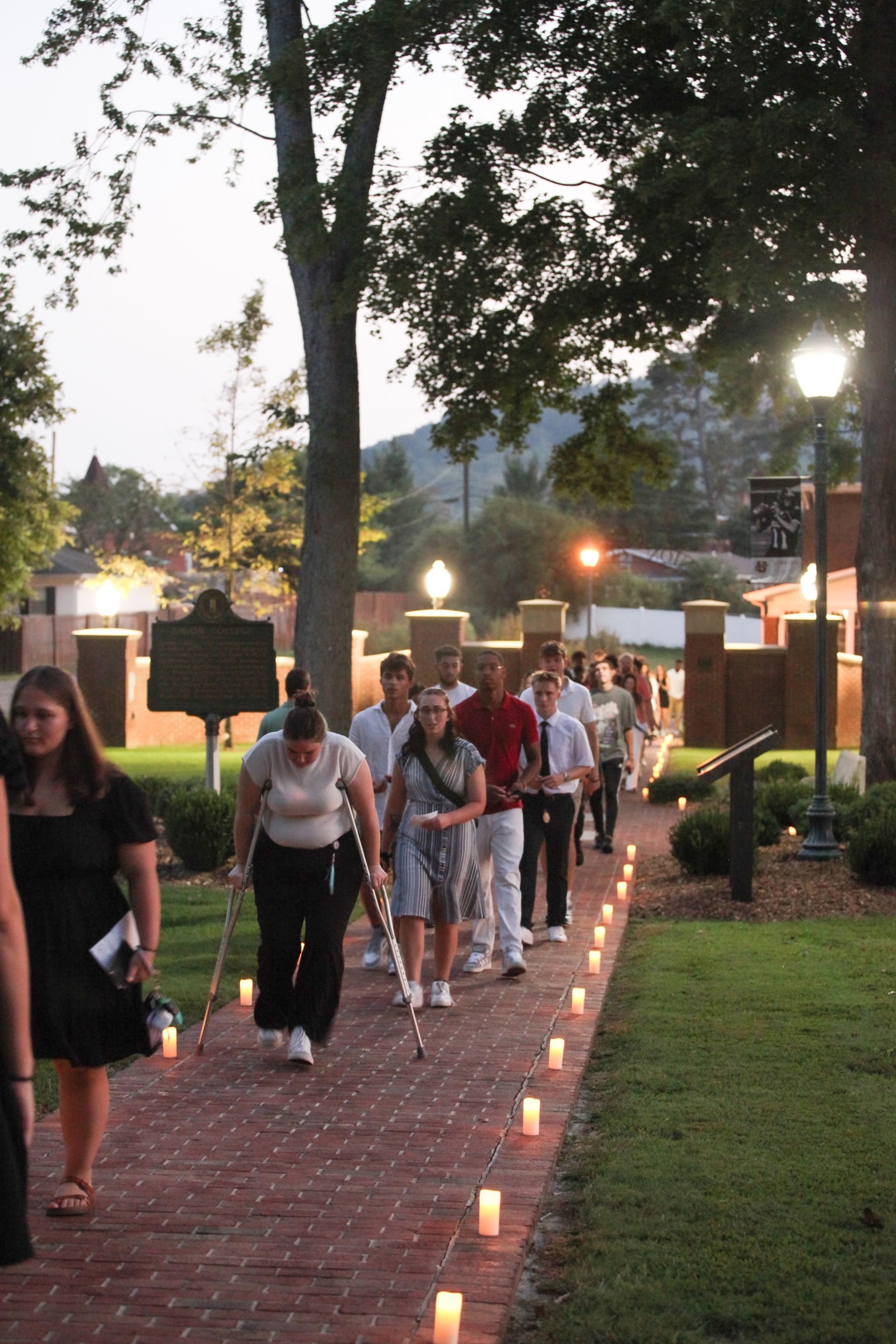 Freshmen dressed in both casual and business-casual attire are being led along the front of campus in the dim evening light. They are walking through the center of the luminary-lit, brick-lined Legacy Walk and into the inner circle. One female student is walking with crutches.