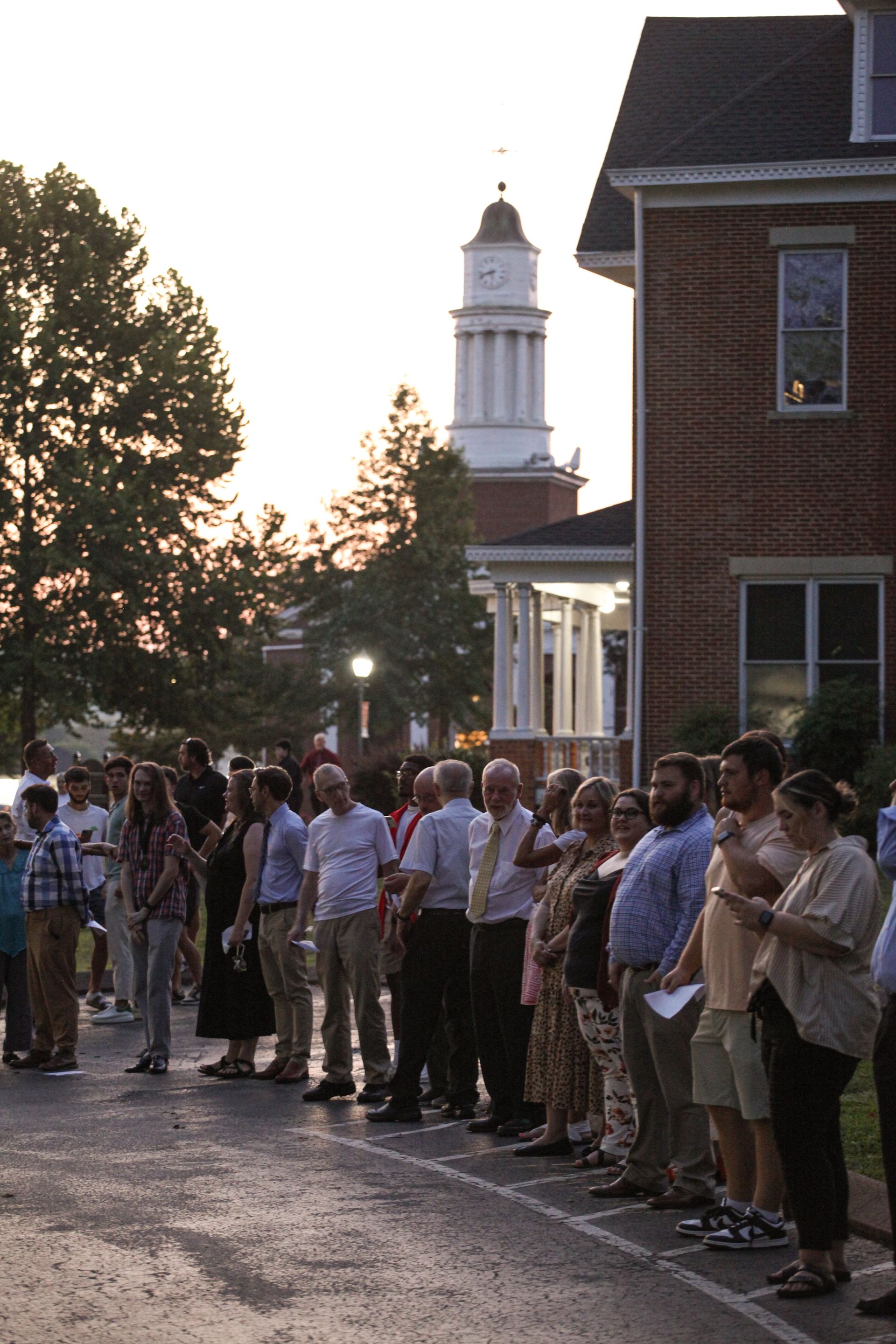 Students, faculty, staff, and community members are lined up in the dim evening light along the outer circle of the luminary-lit Legacy Walk, with Speed Hall and the Conway Boatman Chapel visible in the background. All are wearing a mix of casual and business casual-attire.