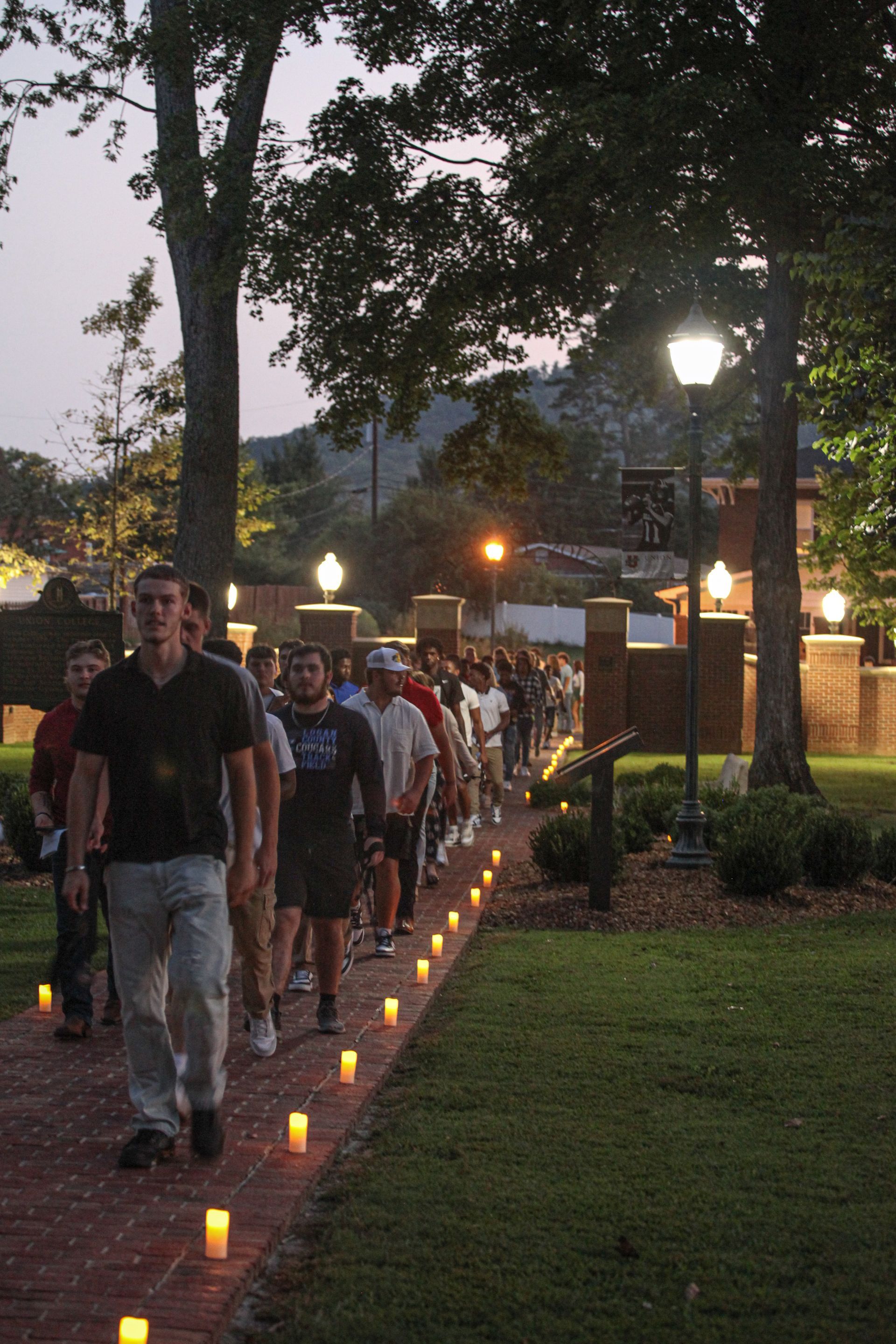 Freshmen dressed in both casual and business-casual attire are being led along the front of campus in the dim evening light. They are walking through the center of the luminary-lit, brick-lined Legacy Walk and into the inner circle.