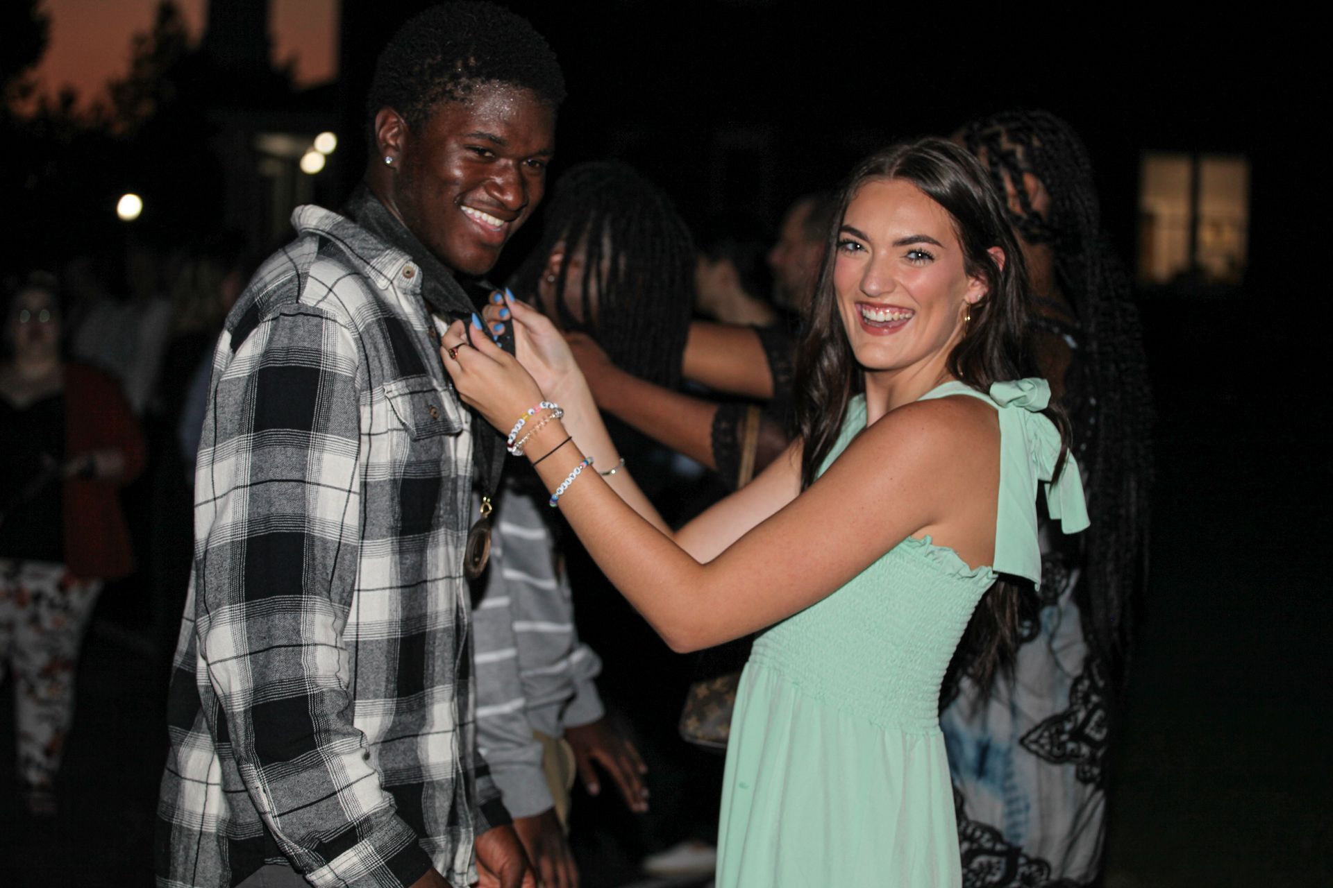 A male freshman wearing a gray checkered shirt is receiving a Convocation Circles medallion from a woman wearing a teal dress. Both are smiling at the camera while she places the medal around his neck. In the background, several other students are also receiving medallions against a dark sky lit by streetlamps.