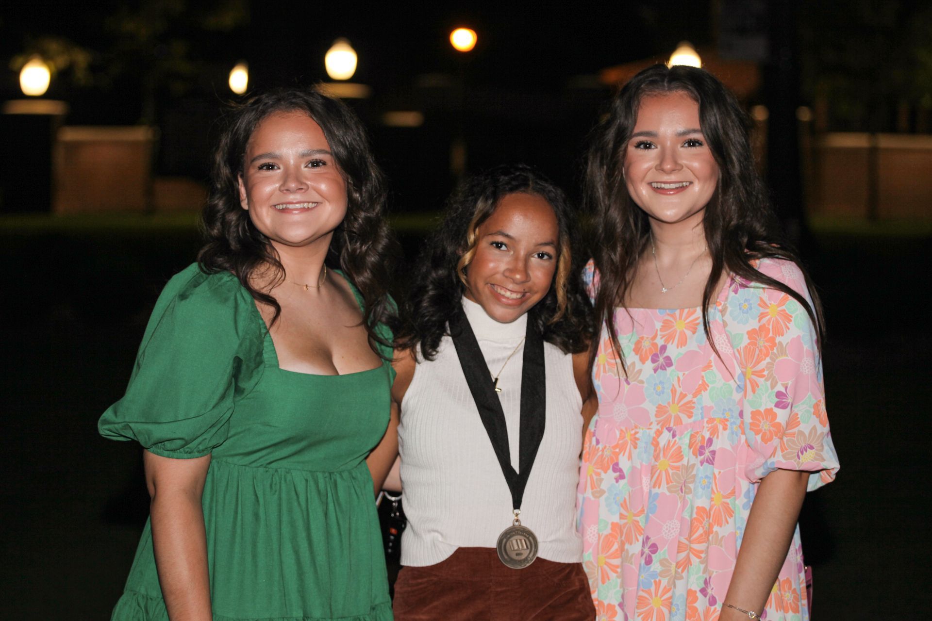 Three female students dressed in formal attire are posing for a photo with their arms around each other. The student in the middle is wearing a Convocation medallion on a black ribbon around her neck, like a necklace. A dark sky and lamplights from the front of the campus are visible in the background.