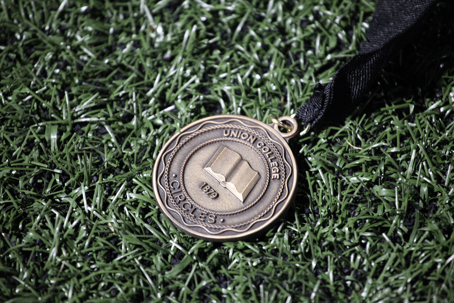A Convocation Circles medal with a black neck ribbon is lying on green grass lit up by soft lighting. The medal reads Union College 1879 CIRCLES.