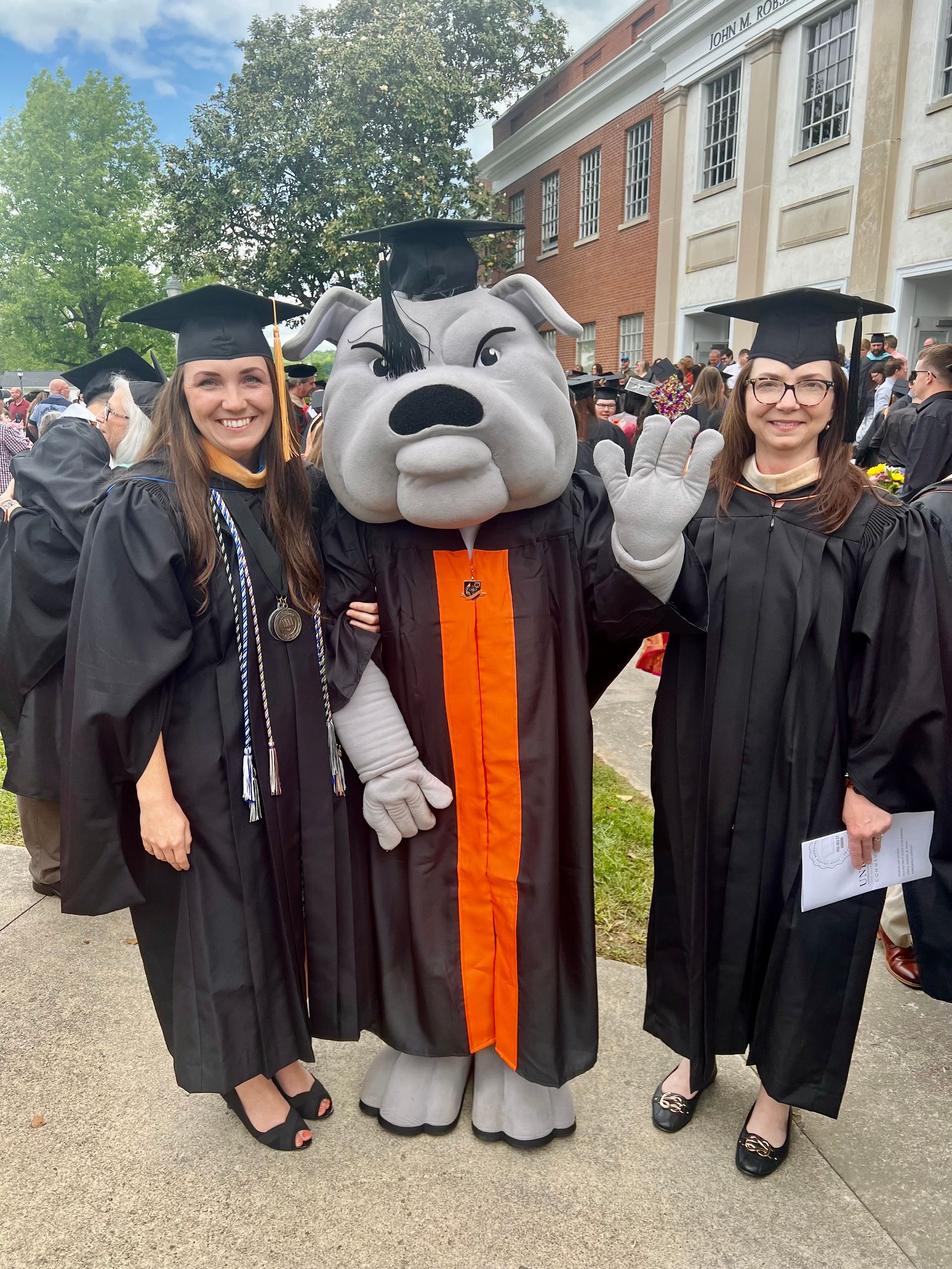 Lynn and Amanda with Union's mascot, Mack the Bulldog, all wearing caps and gowns on Union's campus.