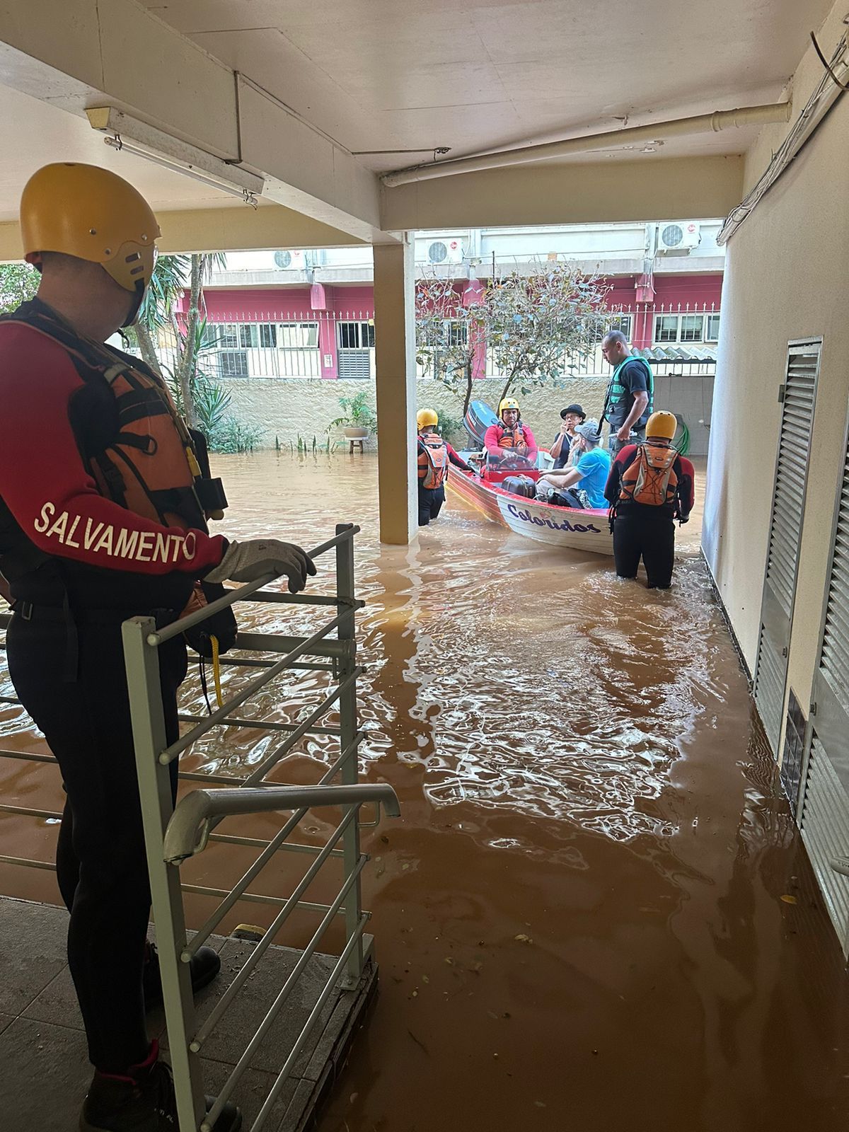 Rescue workers in yellow helmets and bright orange safety gear walk a small boat full of civilians through muddy water as another rescue worker above the water against a railing looks on. They are being brought under a sheltered area, and in the background, a stone wall and red building are visible.