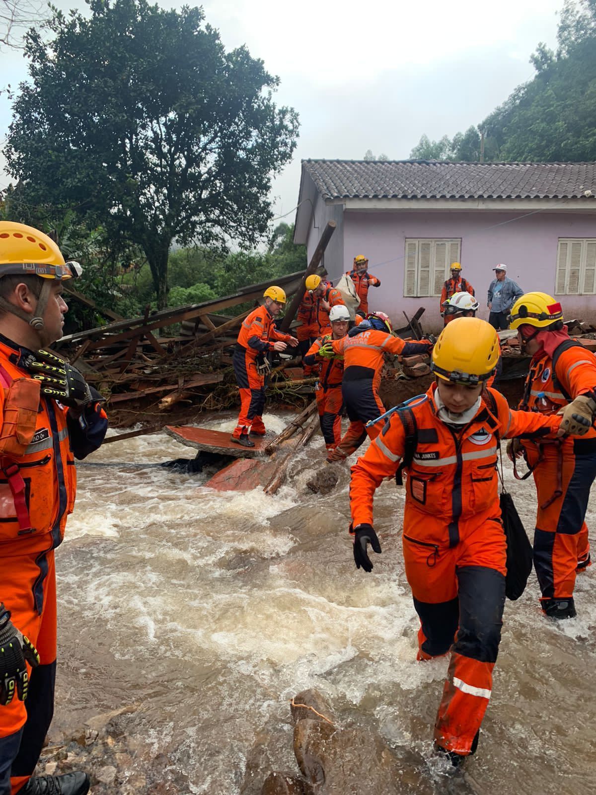 Rescue workers in yellow helmets and bright orange safety gear wade through water and flood debris. A purple house, metal debris, and lush green trees are visible in the background.