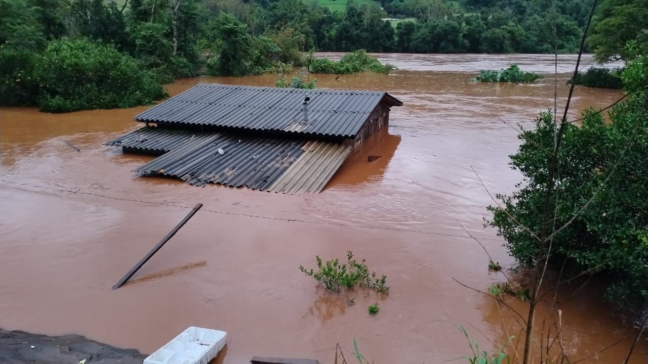 Only the roof of a large building remains above the dark brown floodwater, but part of it looks as though it is starting to submerge. Lush green trees border the floodwater in the background.