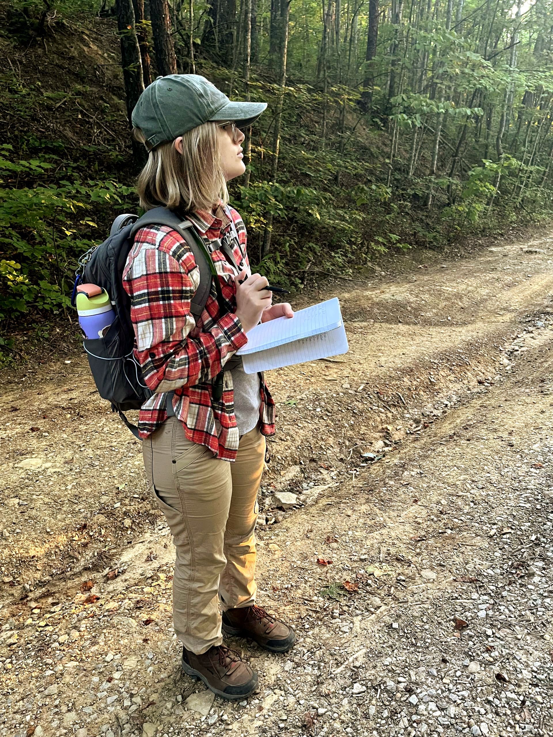 Amelia takes noes in her field journal while standing on a rocky path with forest foliage behind her. She's wearing a backpack, baseball cap, plaid shirt, long pants, and hiking boots.