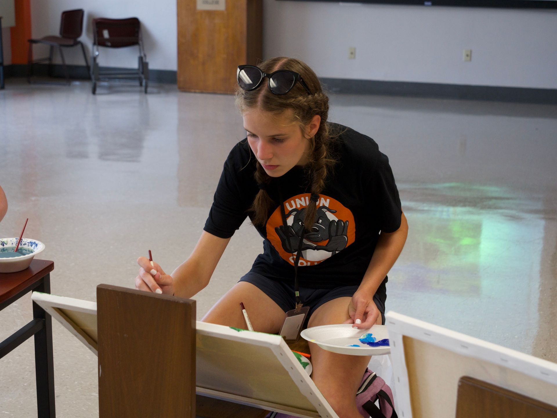A student painting a canvas with a plate of paint in hand.
