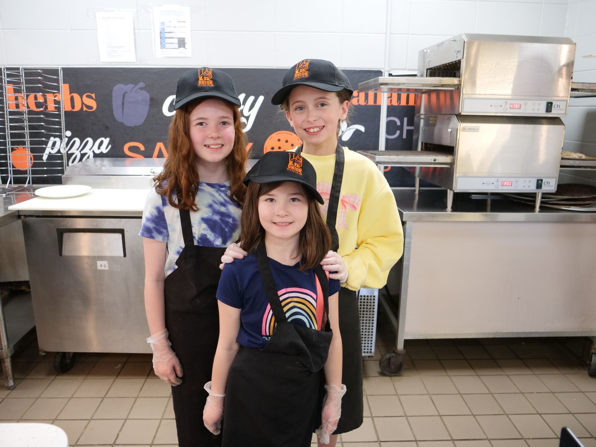 Three kids smiling in Union's kitchen wearing Union Dining hats and aprons.