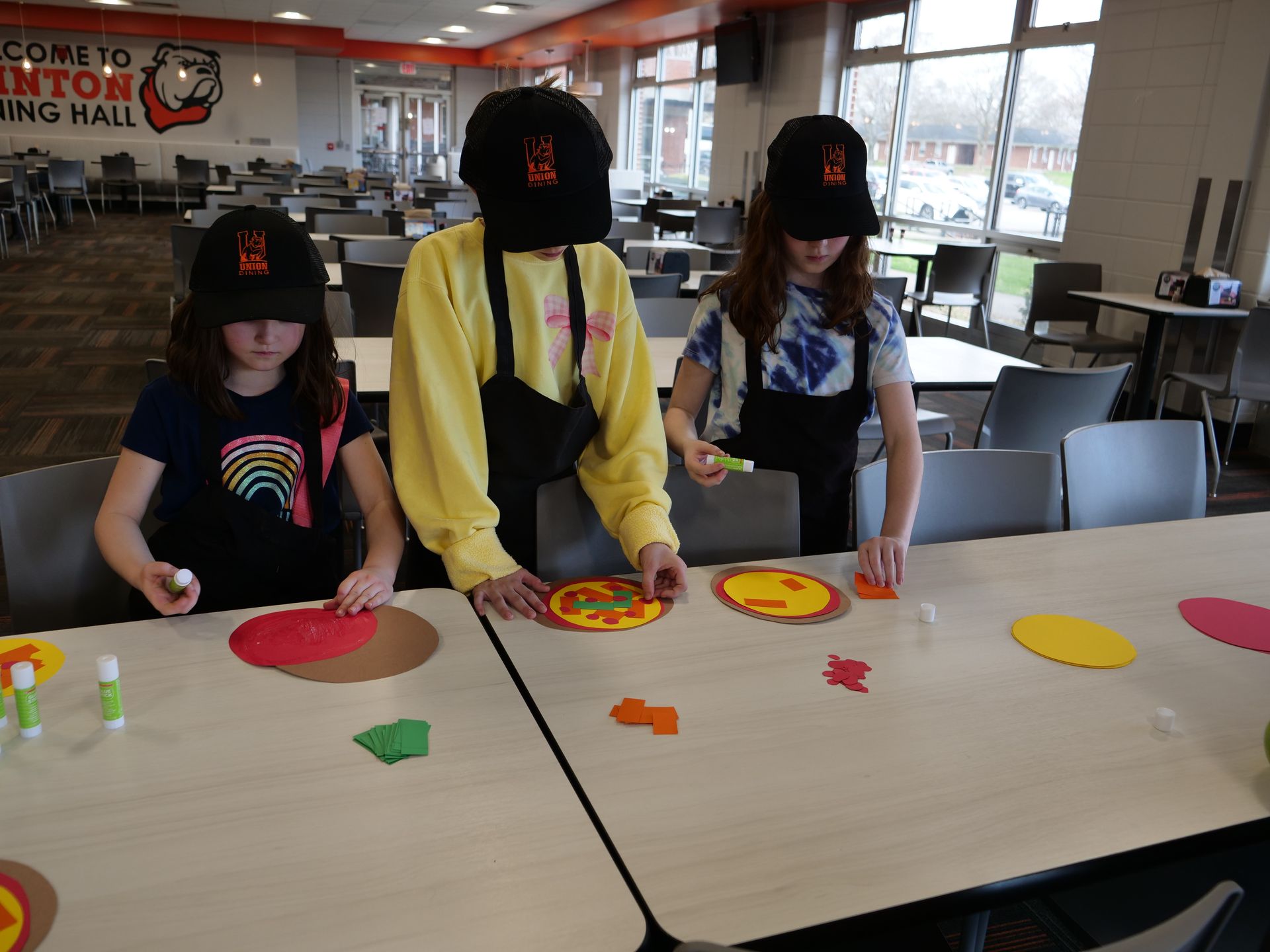 Three kids wearing Union Dining hats and aprons work on decorative paper pizzas.