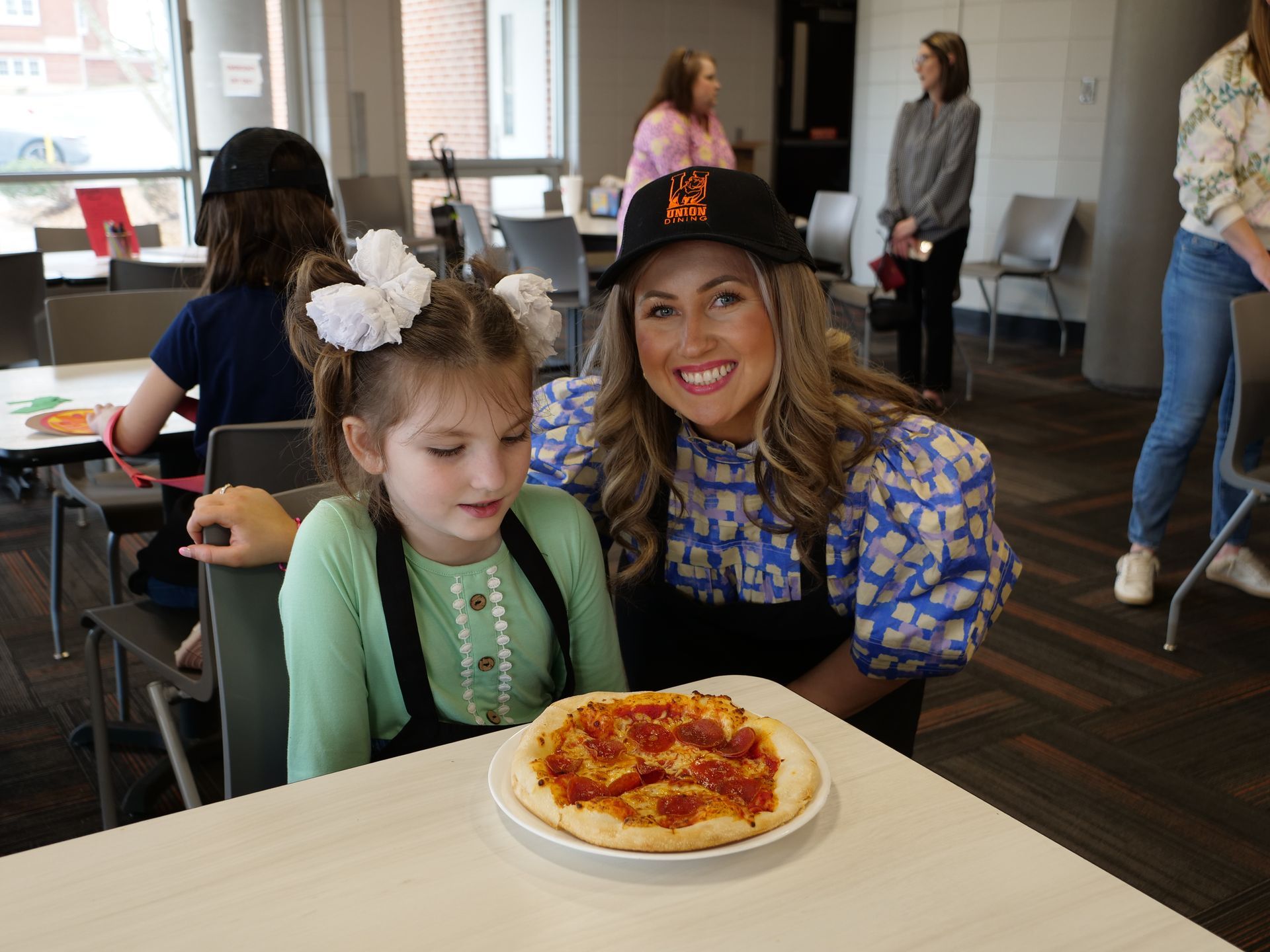 Libby Kate Vaughn and a child attendee smile with a pizza seated at a table corner.