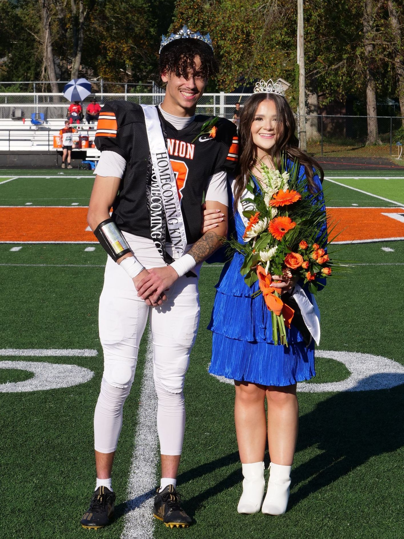 Wearing his football uniform, a silver crown, and a sash that reads HOMECOMING KIND, Tyce smile and leans into Morgan Mays, who is holding his arm with one hand and flowers in the other. She is also wearing a silver crown and sash and is wearing a bright blue dress.