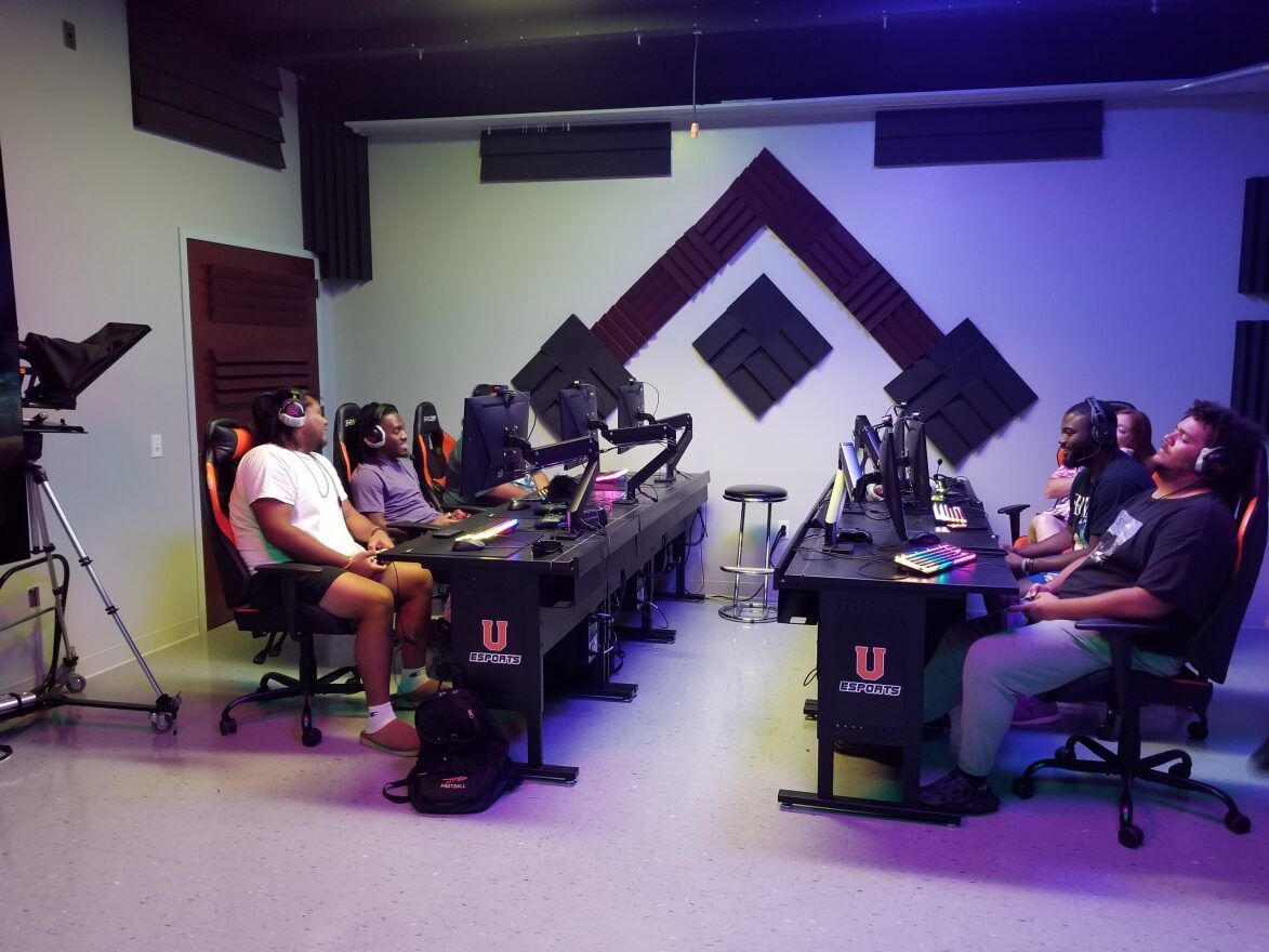 Members of Union's Rainbow Six: Siege team are seated at the PC stations in the Esports Arena, appearing to play video games, talk with each other, and laugh. One has his eyes closed. The black desks the PCs are on have a large orange U logo with the word Esports below it, and the students sit in orange and black gaming chairs.