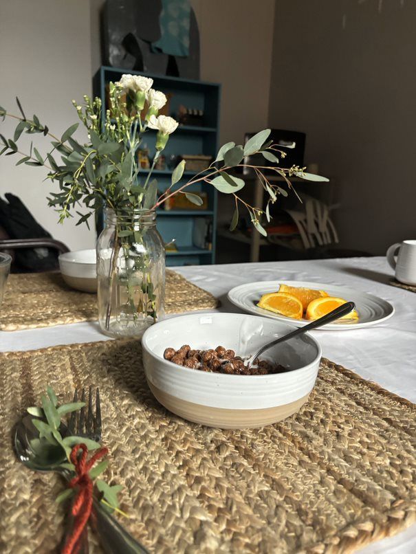 A bowl of cereal and a plate of oranges are lined out on a table. Plants sitting in a glass decorate the center.