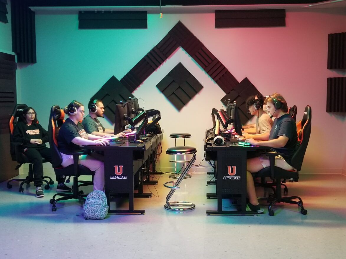 Five members of Union's Valorant team are seated in orange and black gaming chairs at PC stations in the Esports arena wearing headphones and concentrating on the screens in front of them. The black desks the PCs are on feature a large orange U logo with the word Esports below it, and a sixth member sits back a bit, watching them play.