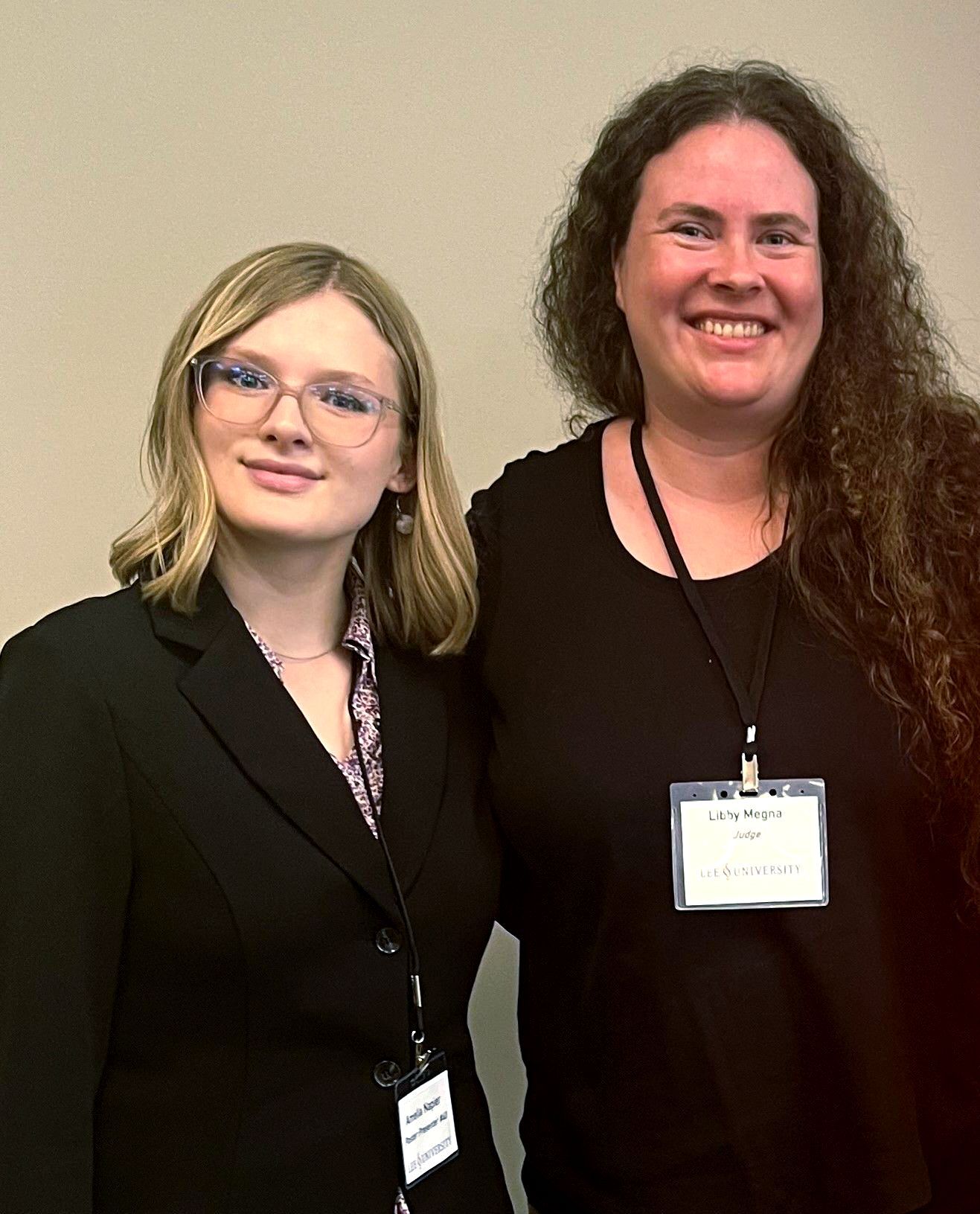 Amelia and Megna pose side by side, smiling and wearing conference badges and black attire.