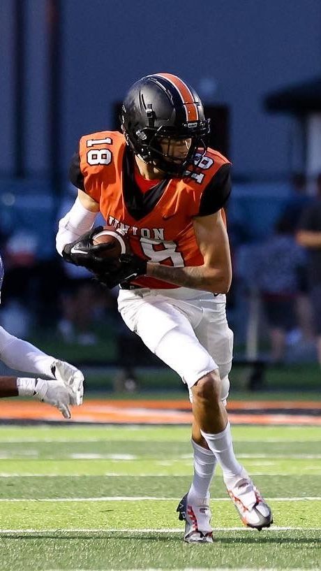 Action shot of Tyce on the football field. He's wearing an orange jersey with while lettering and the number 18, white pants and shoes, and a black helmet. He is clutching the ball and in mid-run.