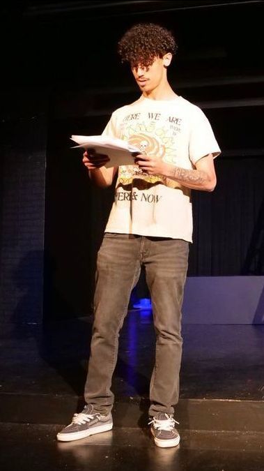 Tyce is a standing on a stage and looking down at a stack of paper he's holding with both hands. He's wearing a t-shirt, black jeans, and sneakers. A large tattoo is visible on his left arm.