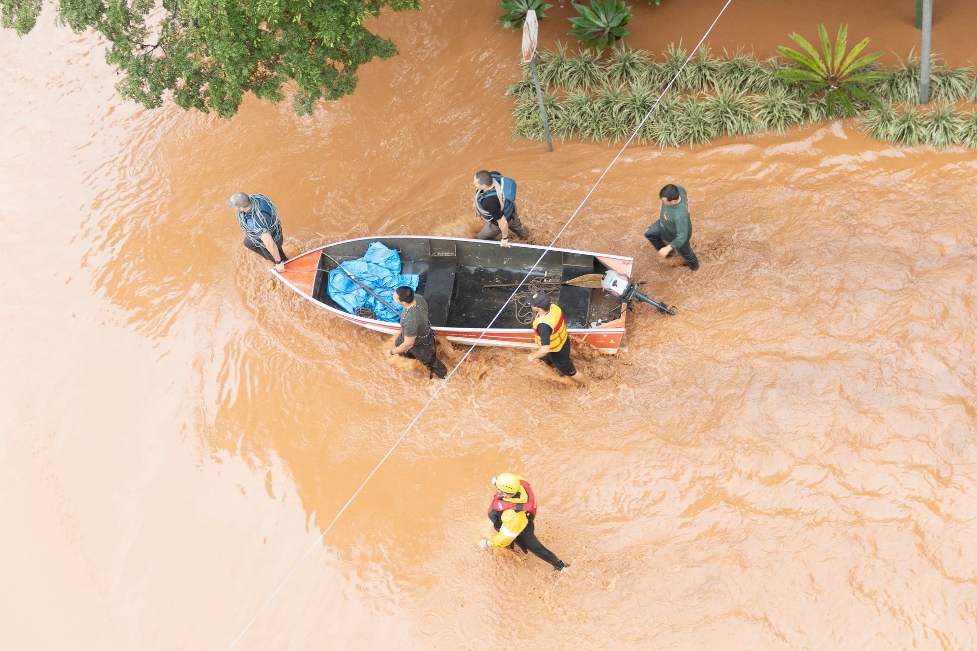 A group of men, some wearing life jackets, wade through bright brown floodwater tugging a boat forward. The boat appears to contain some rescue supplies, and a rescue worker in a yellow helmet and orange life jacket wades several feet to the side of them. Above the boat, a few green plants submerged in water are visible.
