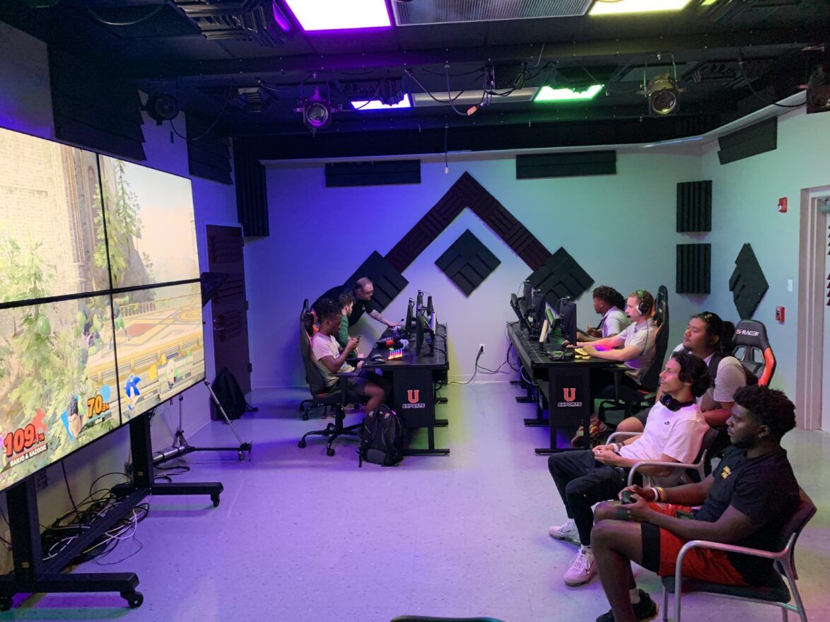 Union students sit playing video games in different areas of the Esports Arena, which is lit with green and purple lighting. Three students near the center play Super Smash Bros. Ultimate on the big four panel screen, while other students in the back play on desktop PCs. Jesse Barton assists a student in the back left.
