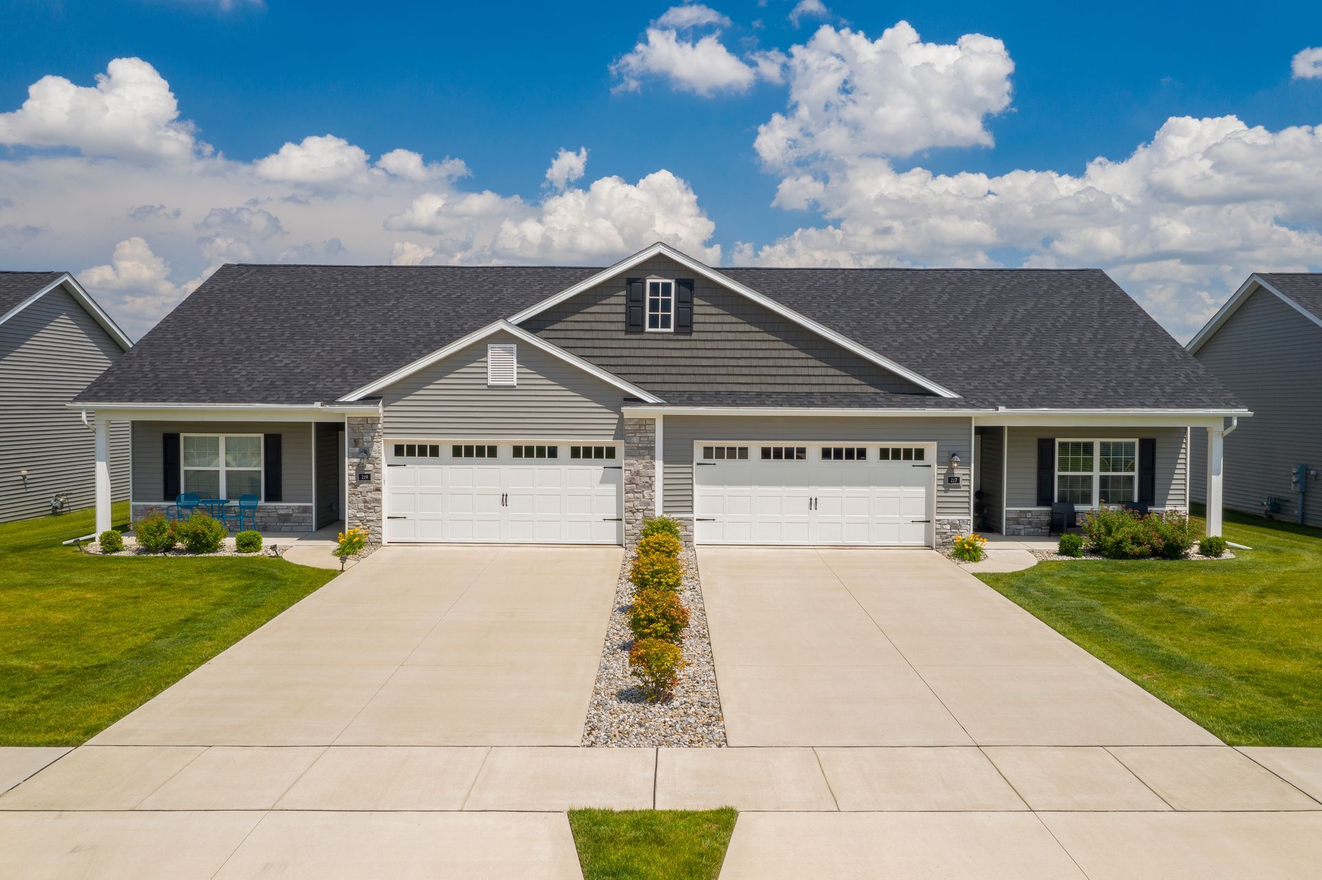 Two-unit, gray duplex with white garage doors and stone accents, blue sky, and green lawn.