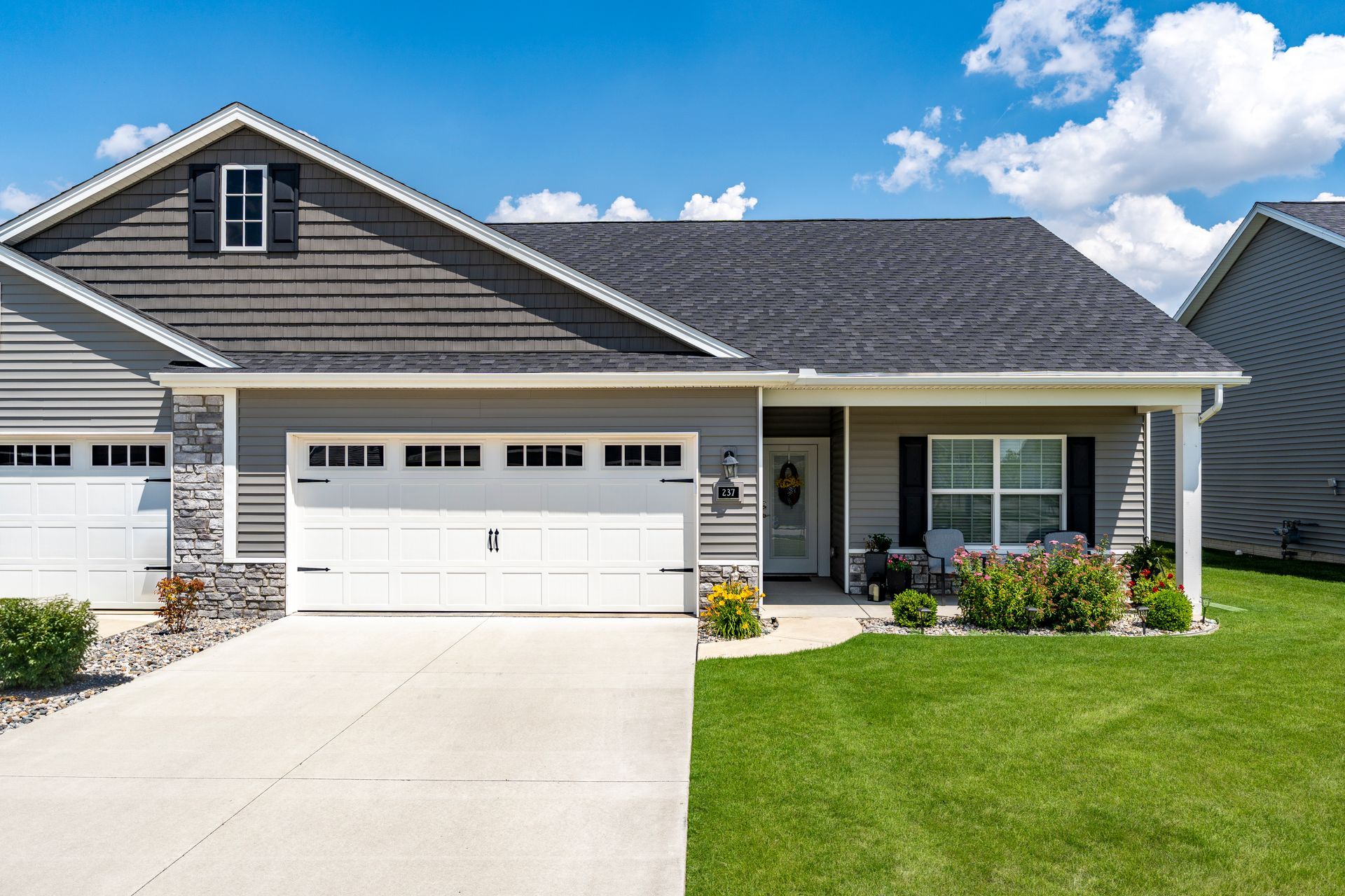 Gray and white suburban house with a garage, front porch, and green lawn under a blue sky.