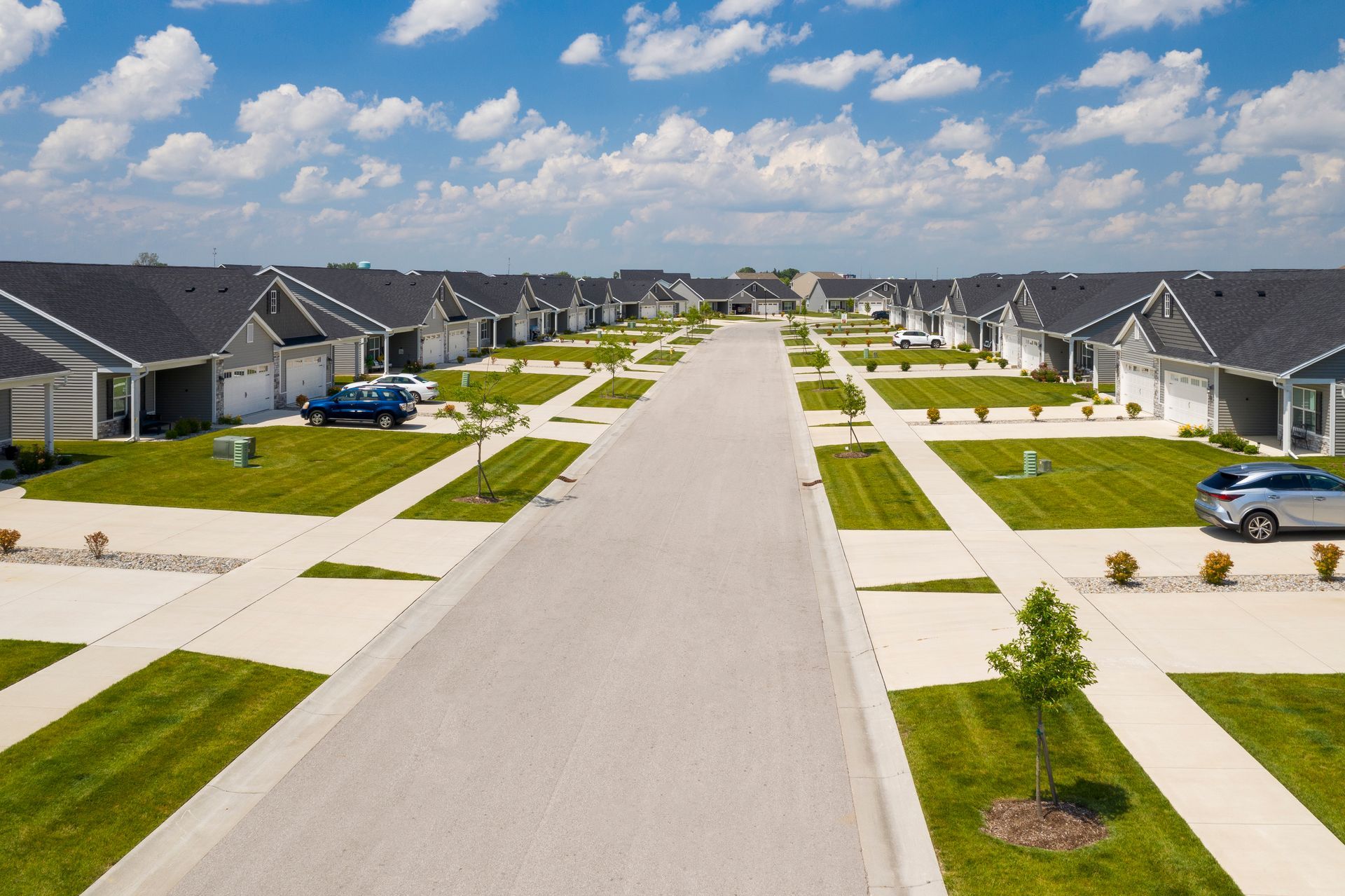 Aerial view of a suburban street lined with matching houses, green lawns, and a blue sky.