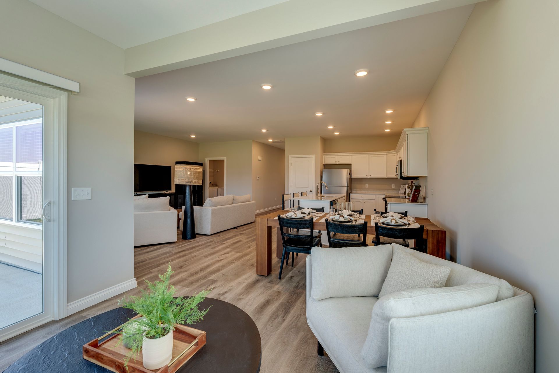 Open-concept living space with kitchen, dining area, and living room visible. Neutral colors, natural light, wood flooring.
