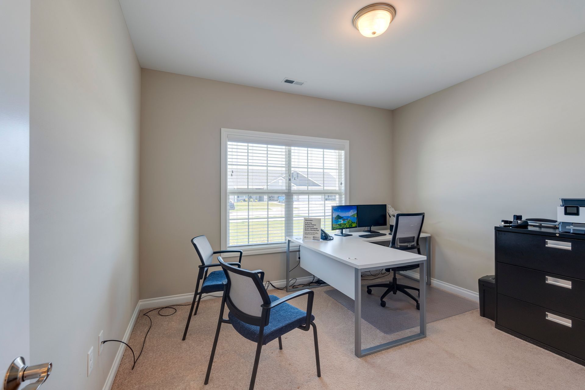 Office with desk, chairs, window, computer, and file cabinet. Beige walls and carpet.