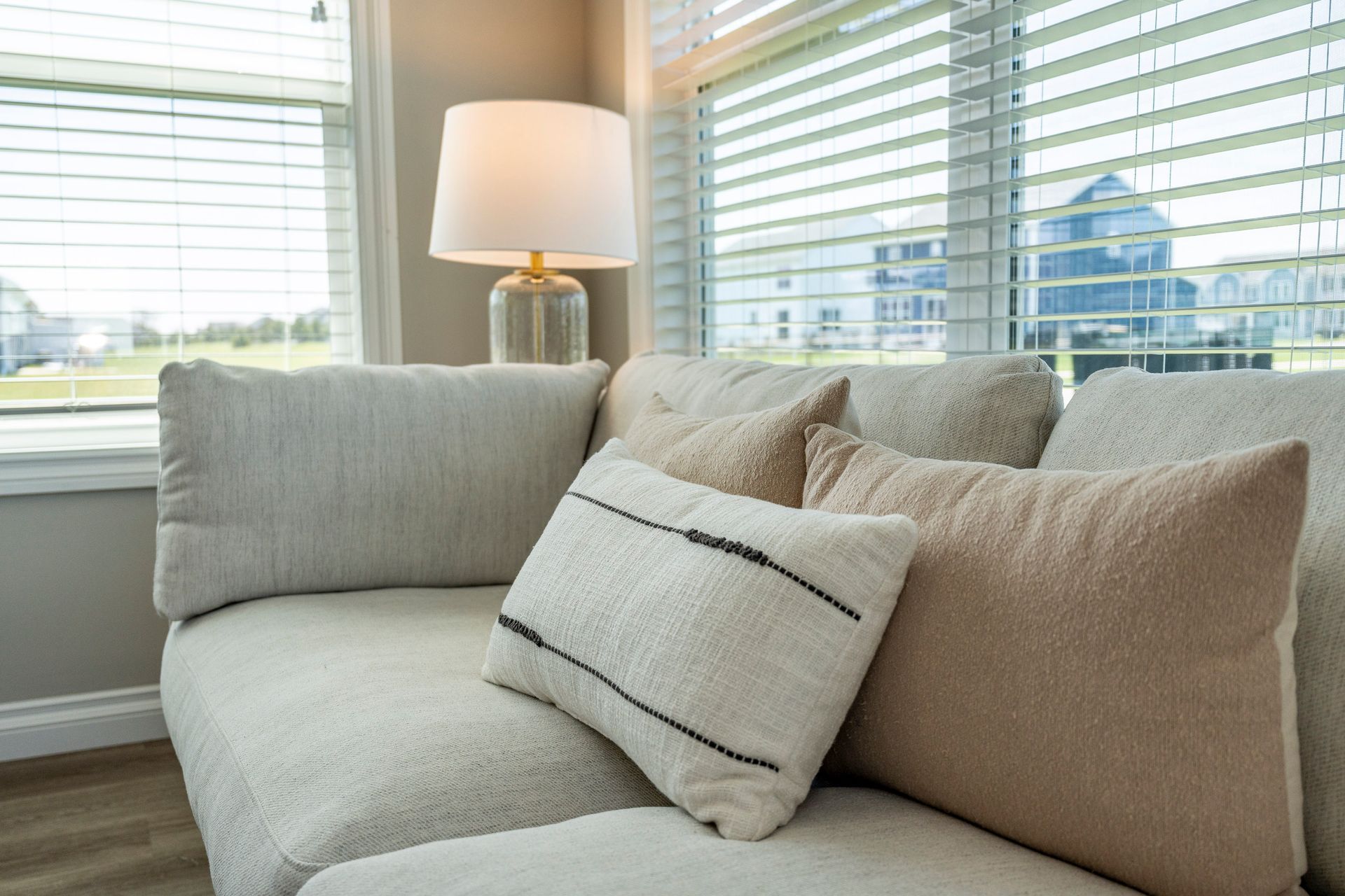Cozy light-colored sofa with pillows near a window, a lit lamp on a table, and a view of the neighborhood.