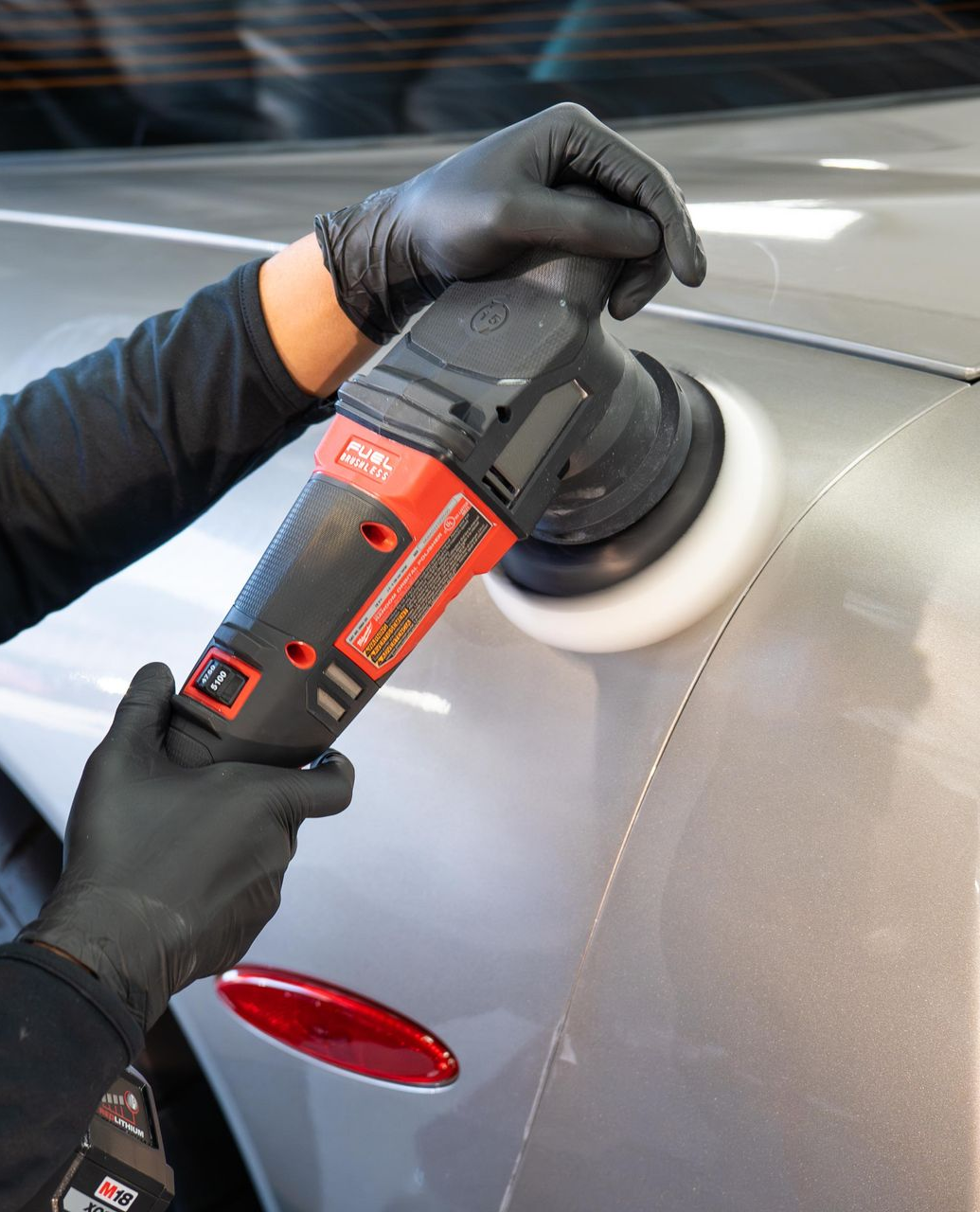 Person polishing a silver car with a red and black electric buffer, wearing black gloves.