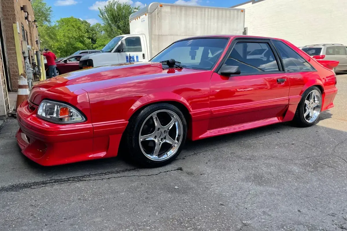 Red Ford Mustang parked on asphalt, custom wheels, bright red paint, sunny day.