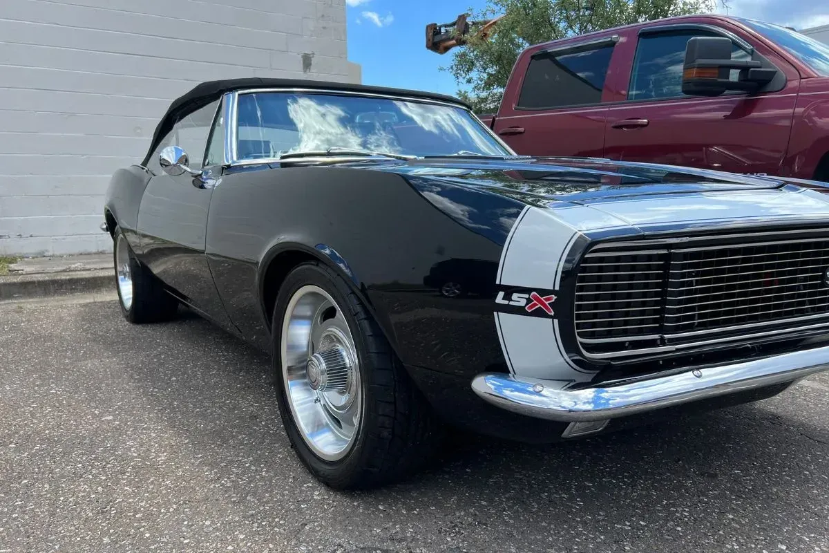 Black convertible Camaro with white racing stripes and chrome trim. Parked on pavement next to a red truck.