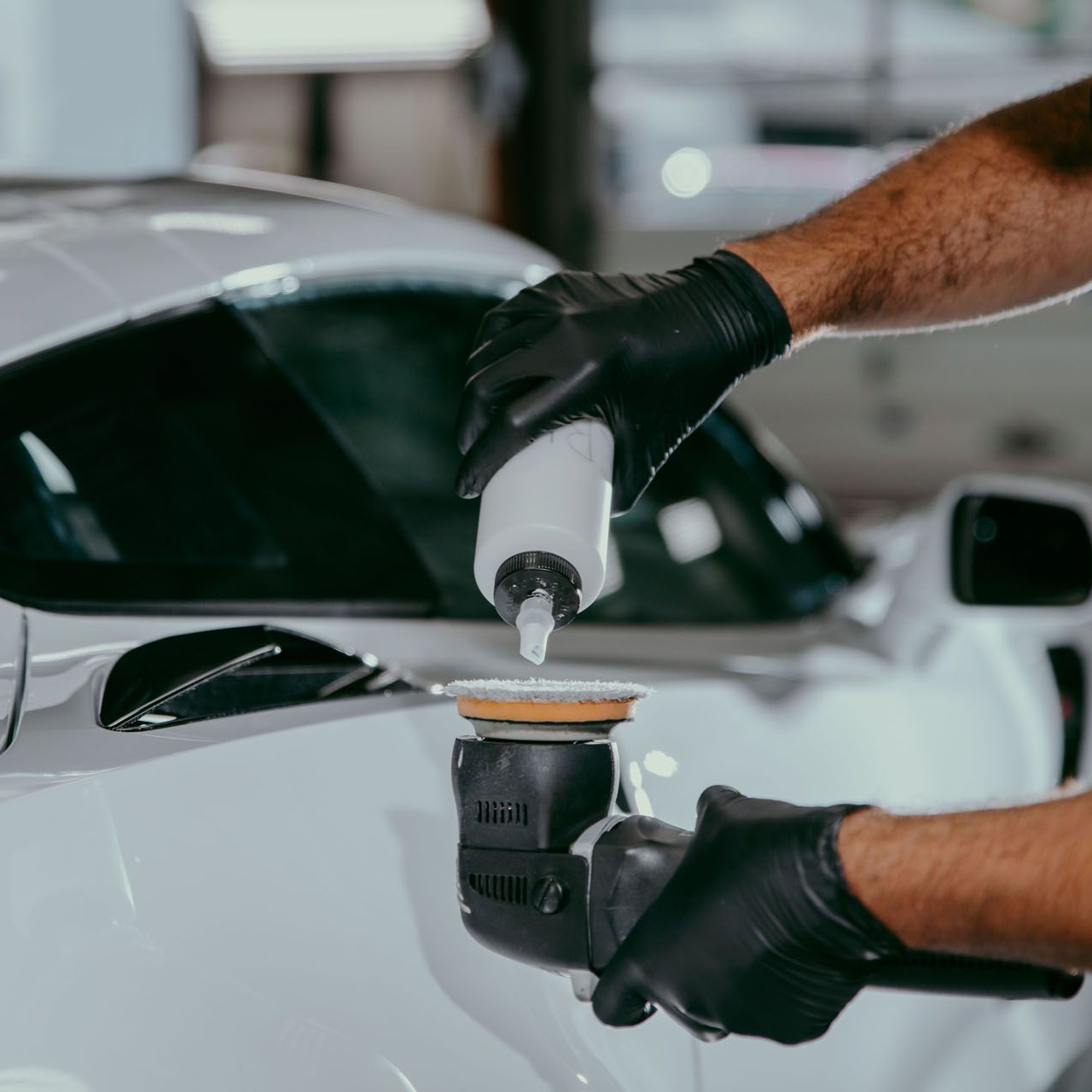 Hands in black gloves applying liquid from a bottle to a polishing pad on a white car.