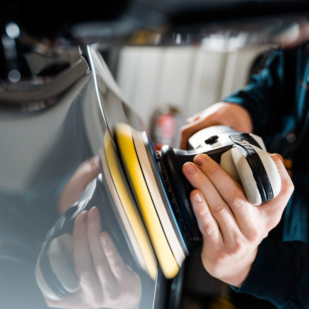 Person polishing car's side panel with a yellow and black electric buffer in a garage setting.